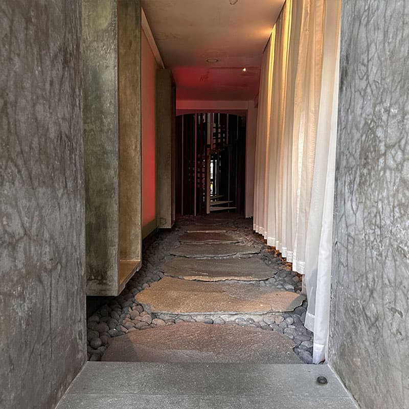 An atmospheric hallway with stone stepping paths, concrete walls, and ambient lighting creating a zen spa-like entrance experience.
