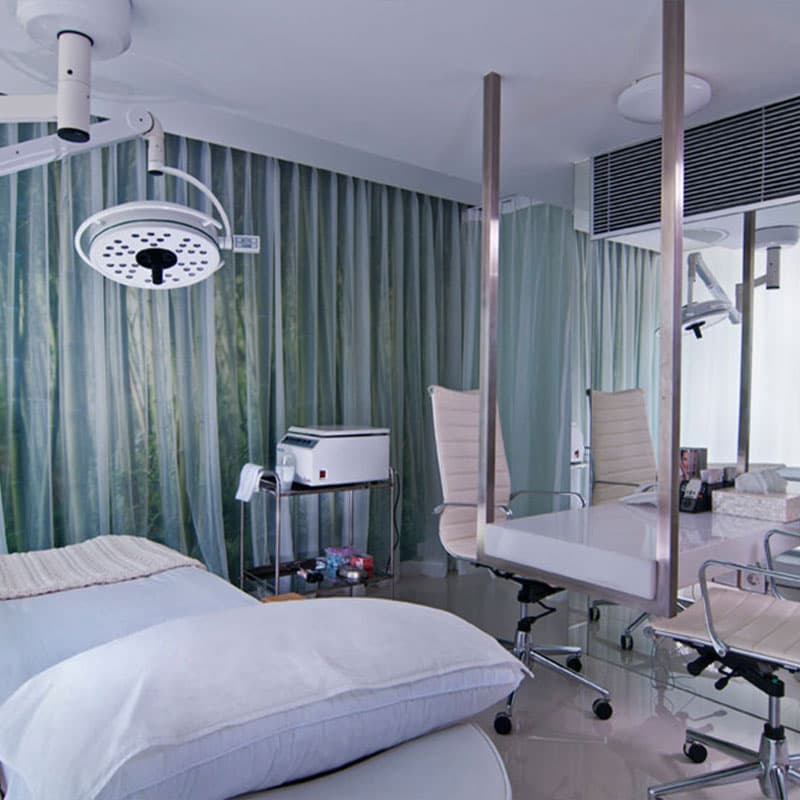A professional and well-lit treatment room with a surgical-grade overhead light, clean treatment bed, modern office chair, and soft green curtain backdrop creating a clinical yet elegant environment.