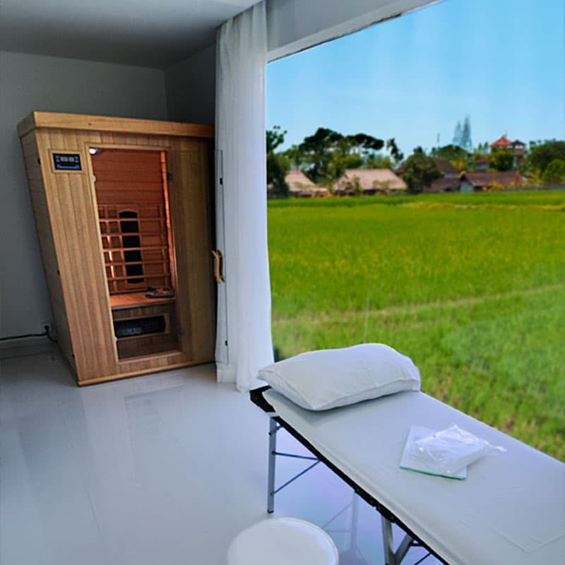 A minimalist treatment room featuring a clean treatment bed and infrared sauna with a spectacular floor-to-ceiling window overlooking lush green rice paddies.