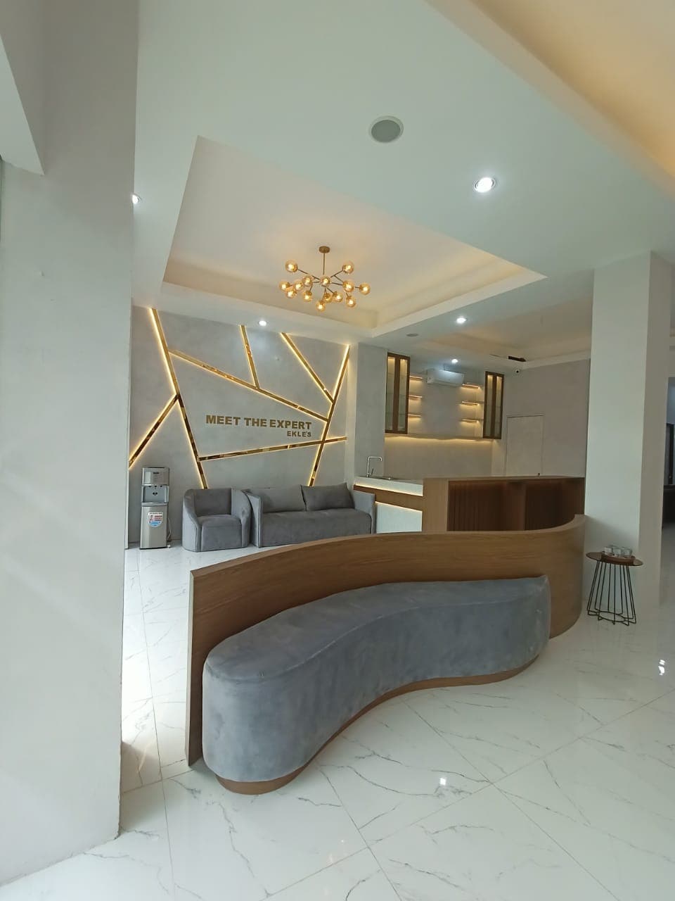 Modern waiting area with Meet The Expert gold signage, geometric gold light strips, curved bench, marble floors.