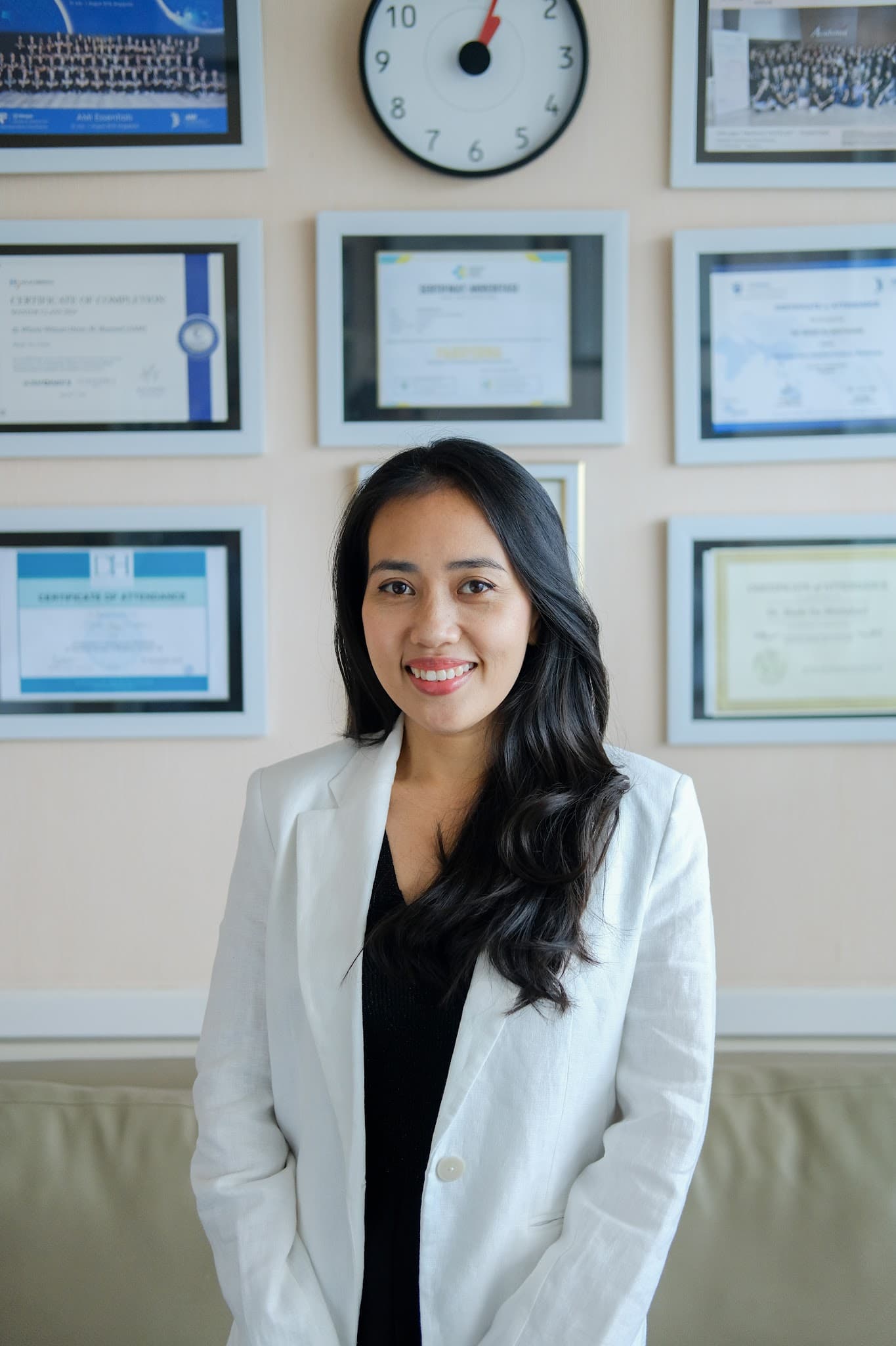 Warm professional portrait of a young female doctor in a white blazer smiling brightly in front of a credential wall with framed certificates. Friendly and welcoming demeanor.