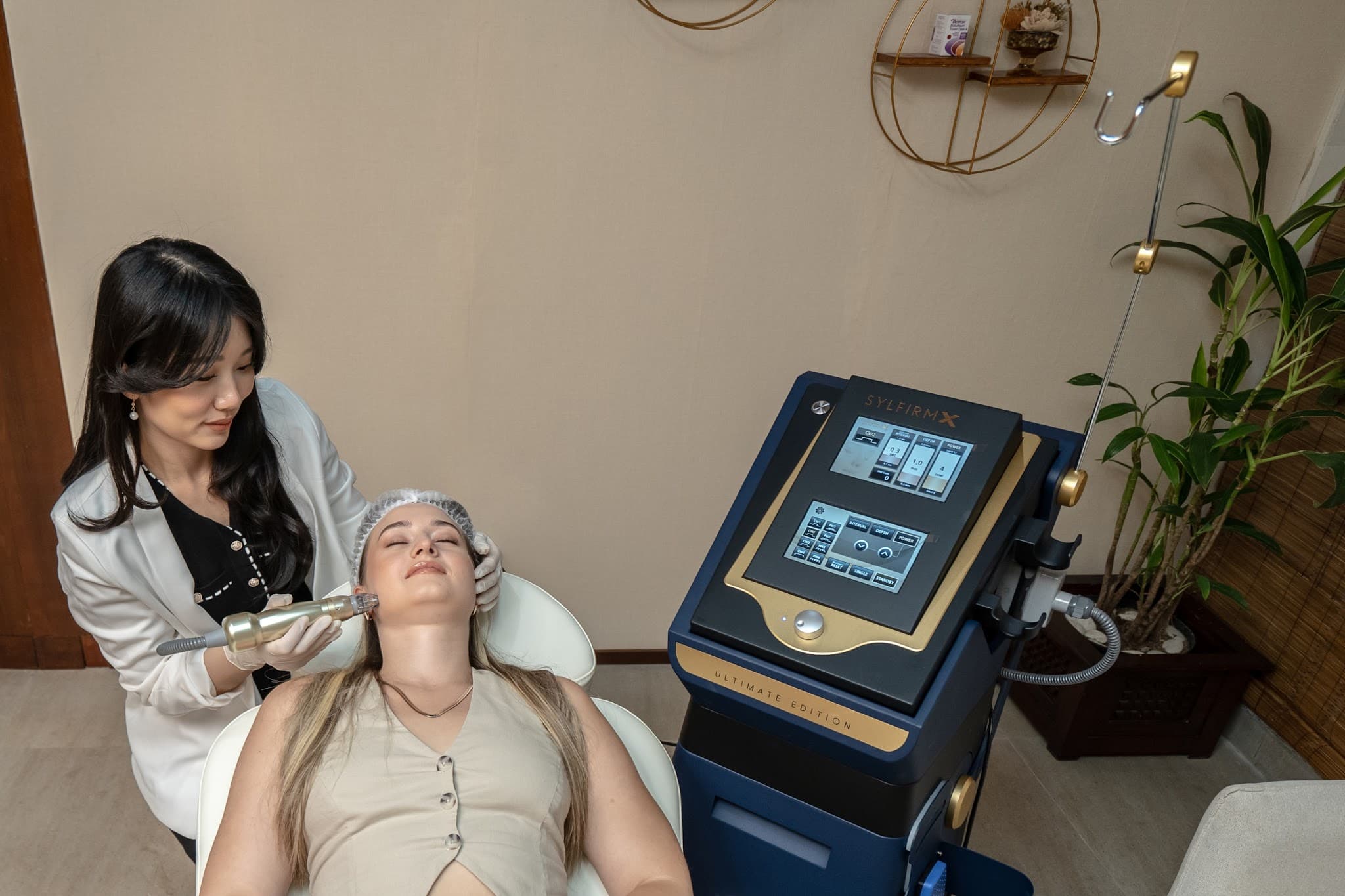 Doctor performing a Sylfirm X microneedling treatment on a client reclined in a treatment chair. The premium navy and gold device is prominently displayed with its screen visible. Clean clinical setting with plants and warm decor.