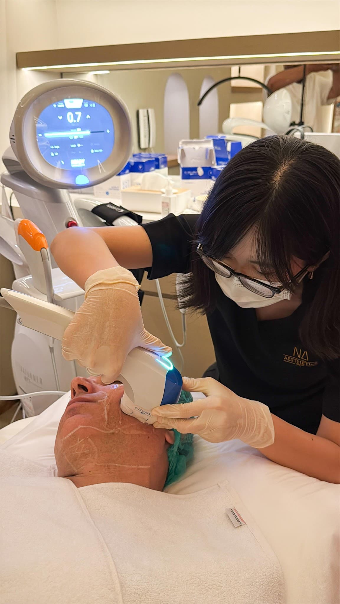 Close-up of practitioner wearing glasses, mask, and gloves performing facial laser treatment on client jawline. Clinical professionalism but cluttered background and close-up angle with treatment markings is borderline.