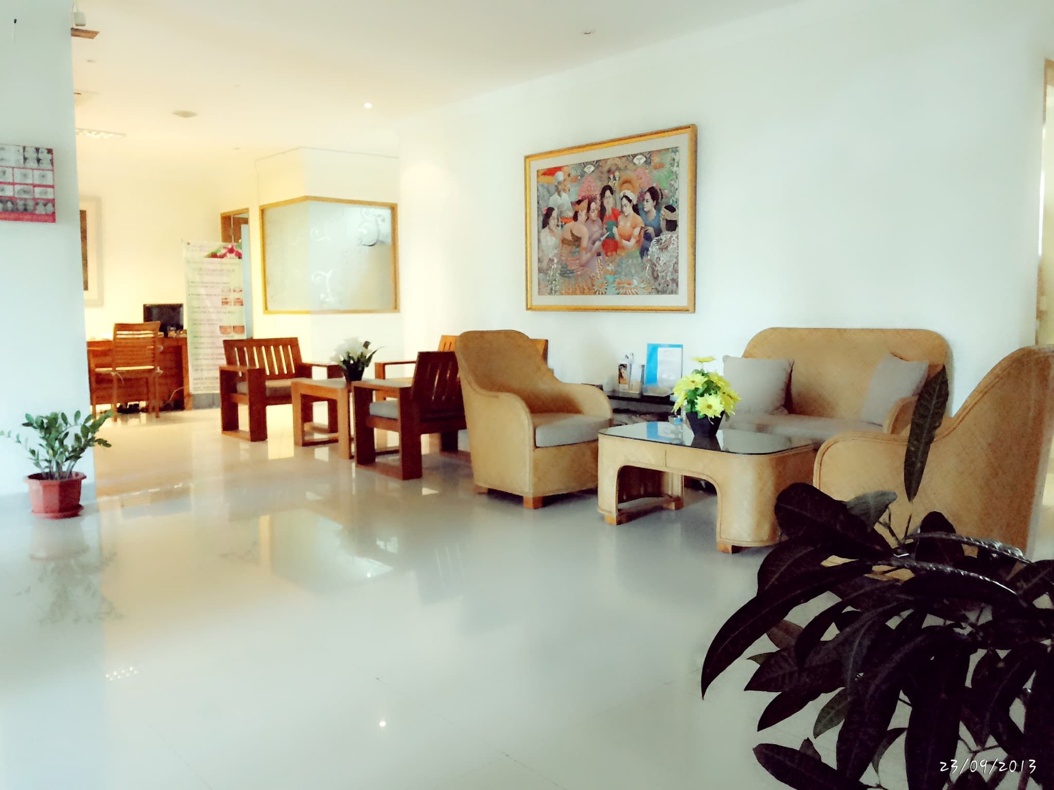 Clinic waiting area with beige sofas, wooden chairs, glass coffee table, artwork, polished floors. Clean but slightly overexposed with date watermark.