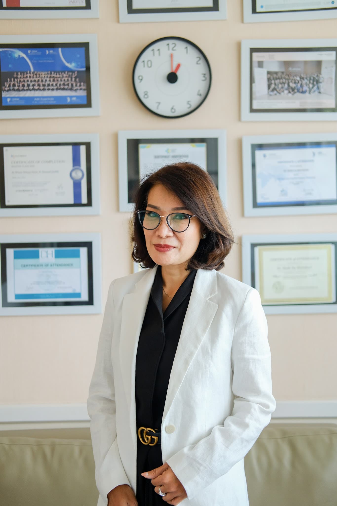 Professional portrait of a female doctor in white blazer and glasses standing in front of a wall of framed certificates and credentials. Confident, approachable expression conveys expertise.