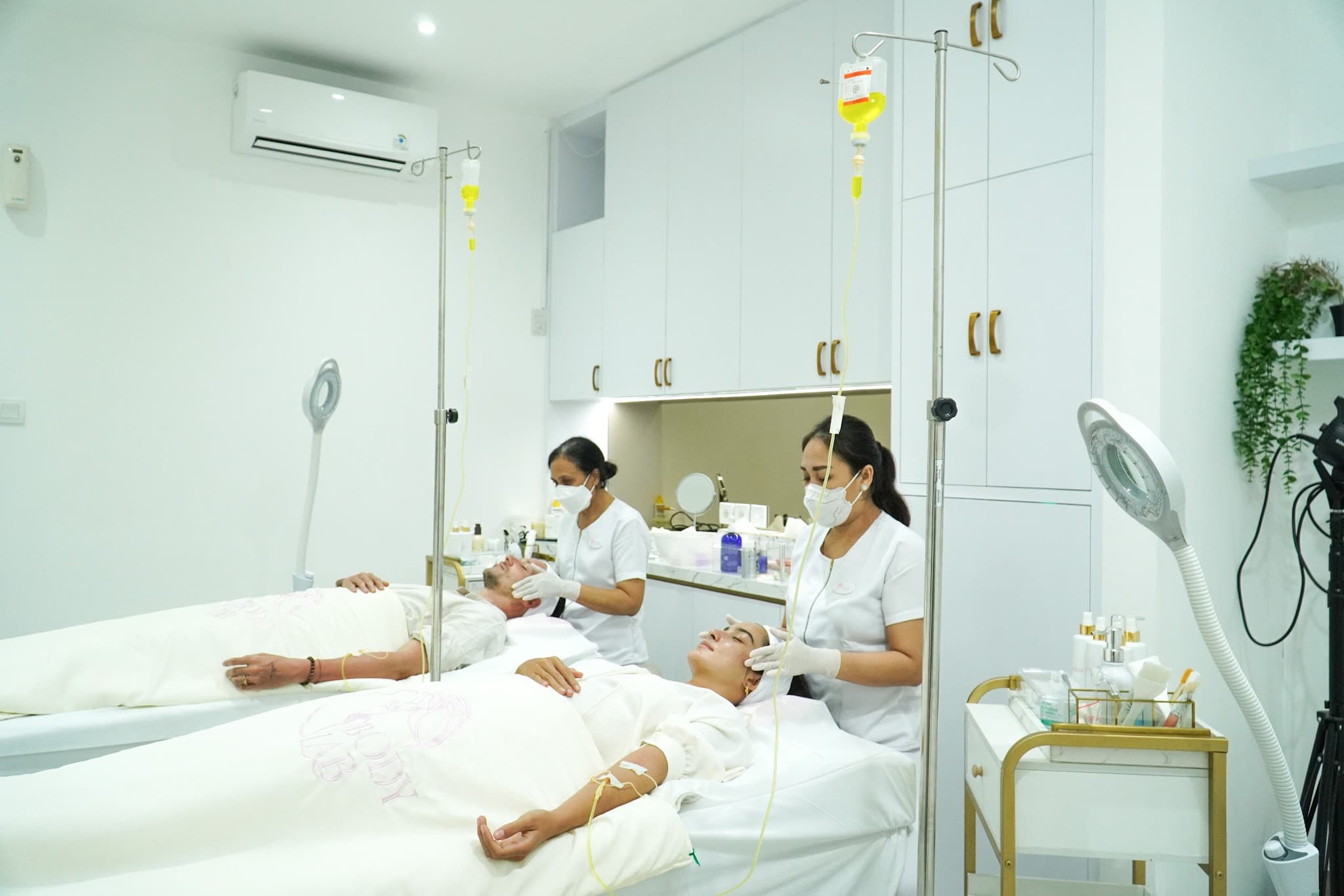 Two clients receiving IV drip therapy and facial treatments from masked practitioners in a pristine white treatment room. Professional clinical environment with gold accent cart, magnifying lamp, and organized product shelving. Conveys professionalism and human warmth.