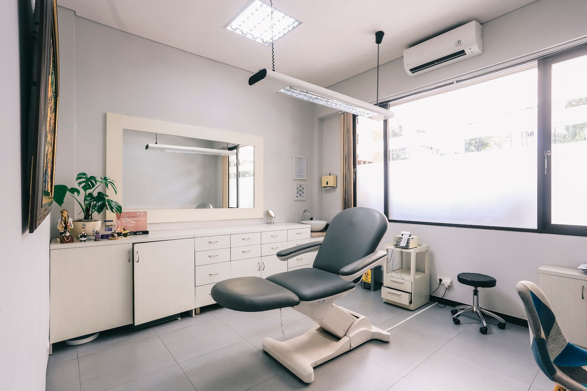 Consultation/treatment room with grey chair, large mirror, white cabinetry, natural light from windows, potted plant, artwork. Clean with good natural light.
