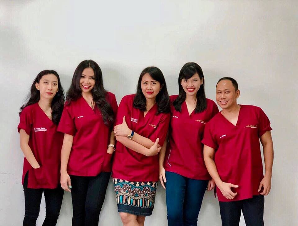 Five team members in matching red/maroon scrubs posing on white wall. Friendly but slightly soft with flat lighting.