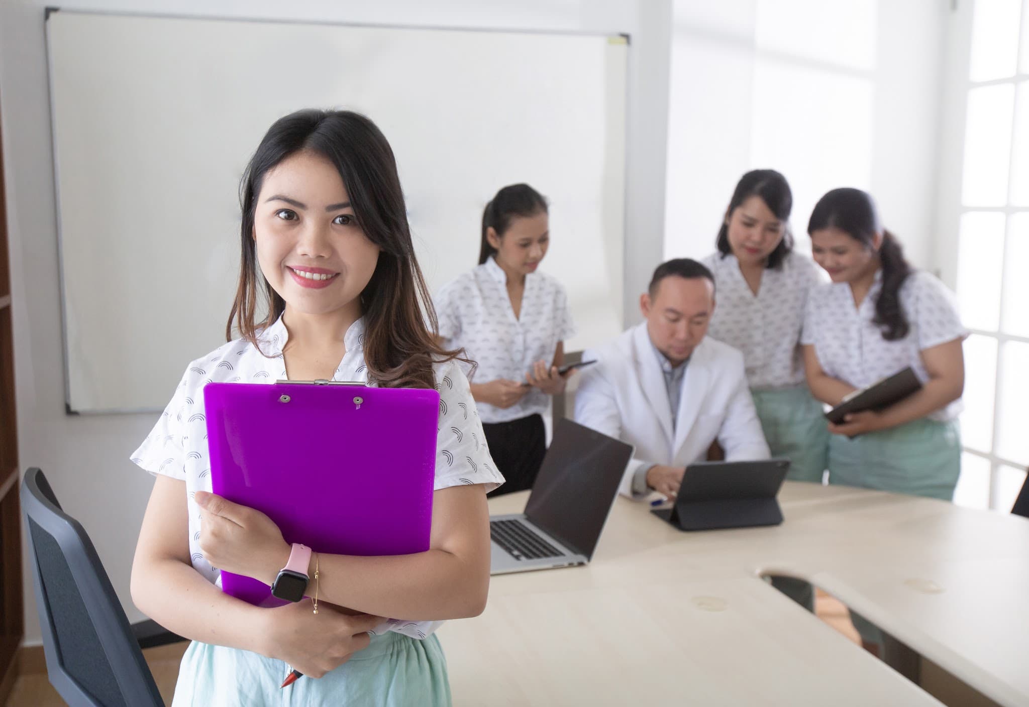 Professional team photo with a smiling staff member in the foreground holding a clipboard, while colleagues and a doctor collaborate at a conference table in the background.