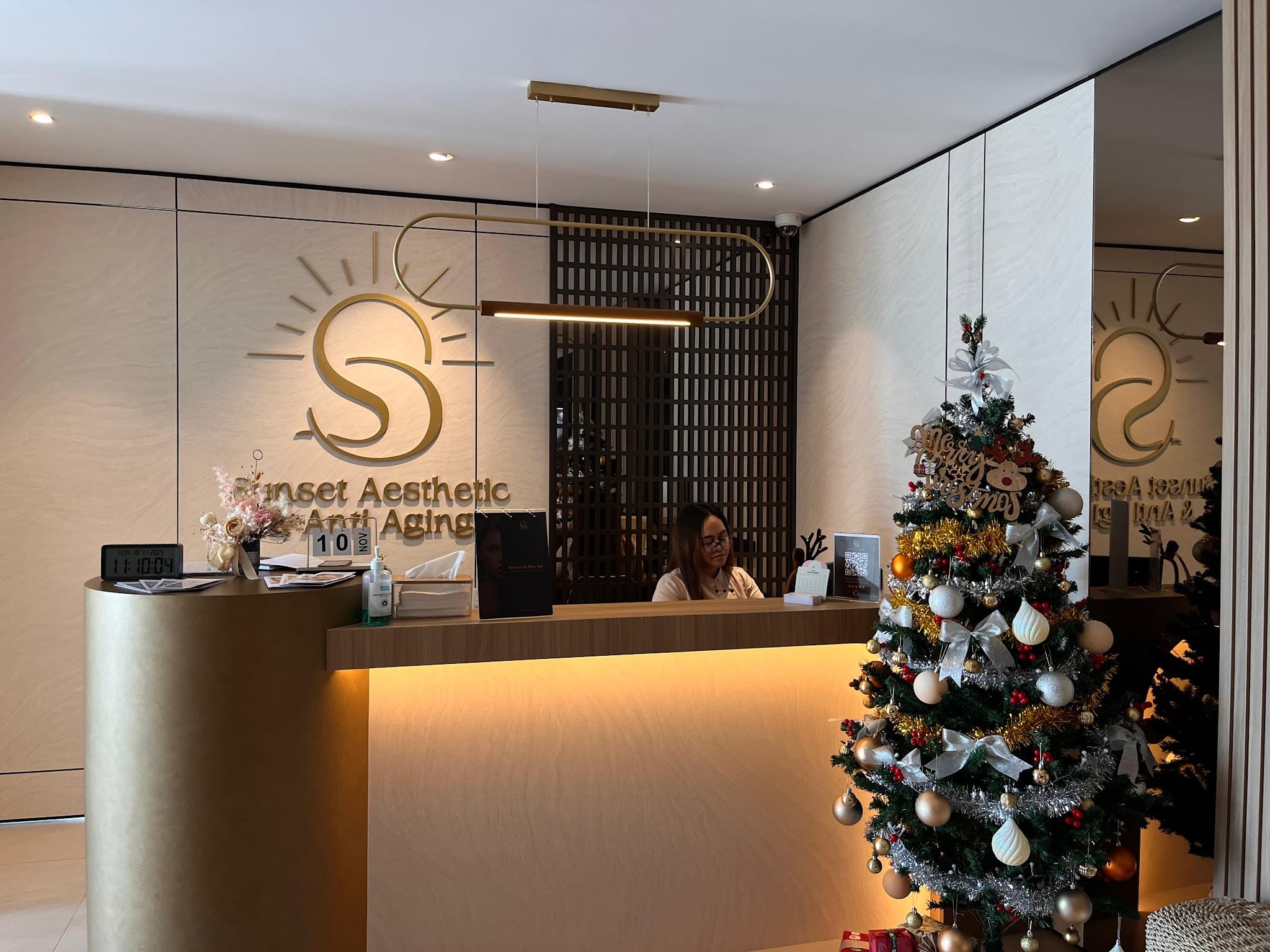 Aesthetic clinic reception area with elegant gold Sunset Aesthetic Anti Aging logo on the wall, a receptionist working behind a sleek backlit counter, warm lighting, and a Christmas tree adding seasonal decor.