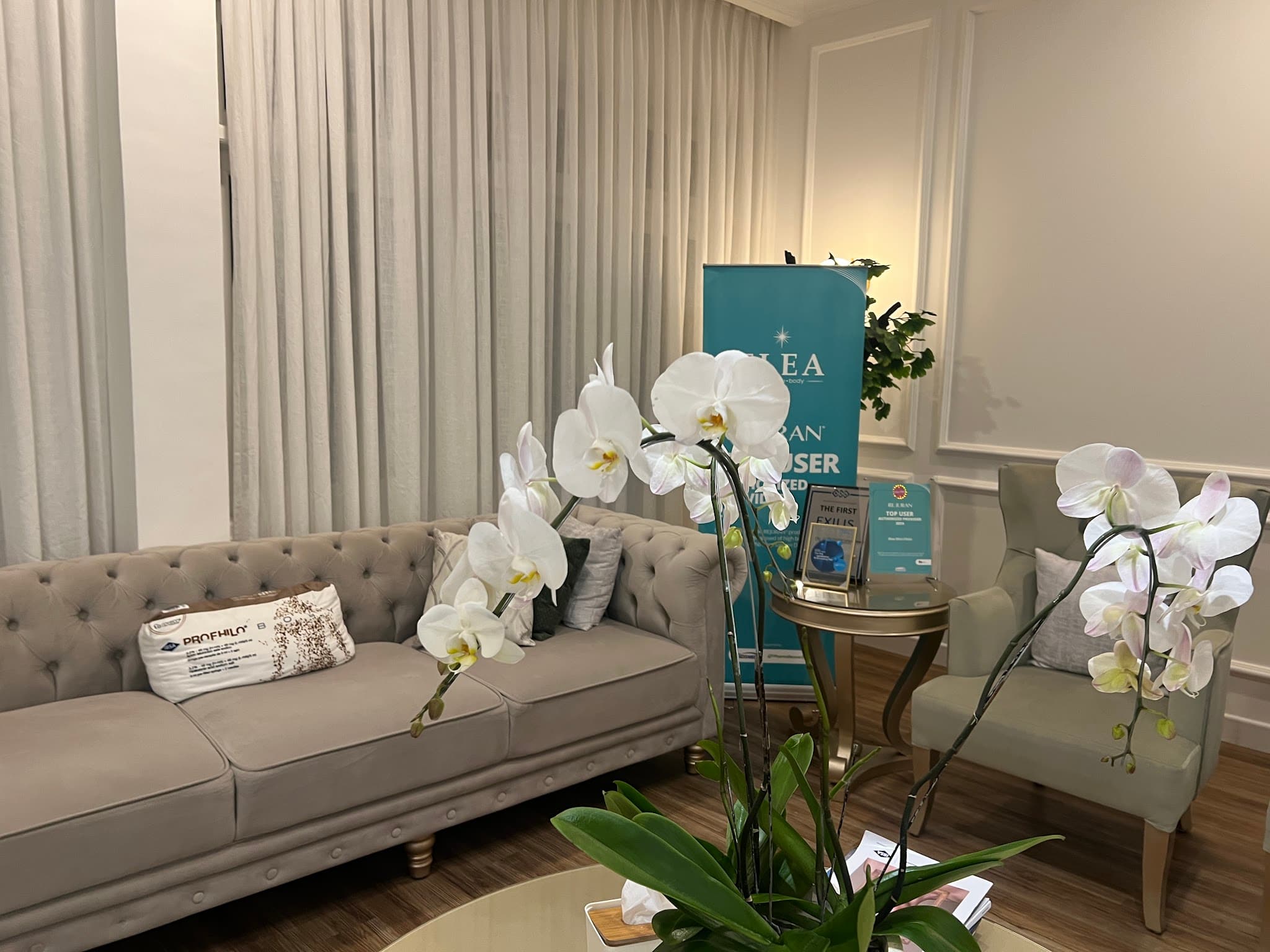 Clinic waiting area featuring a tufted grey sofa, sage wingback chair, and beautiful white orchids in the foreground. A teal promotional banner and branded product pillow on the sofa detract from the otherwise elegant scene. Nice floral elements add warmth.