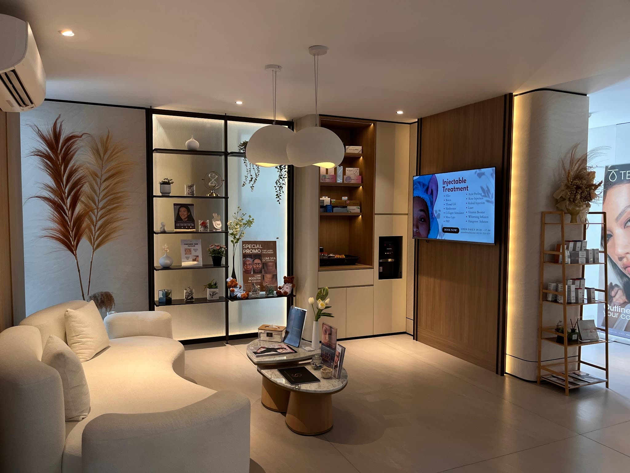 Modern aesthetic clinic waiting lounge with elegant cream curved sofas, marble coffee table, designer pendant light, display shelving with products, and warm wood accents.
