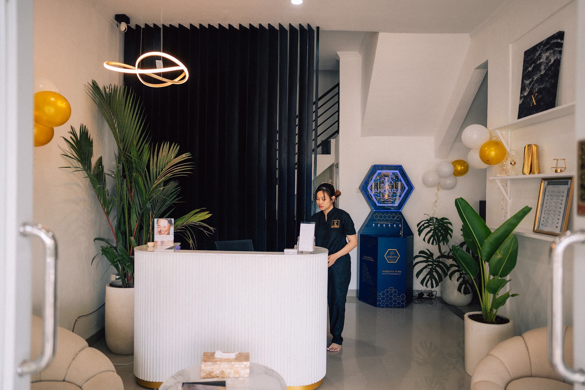 Modern clinic reception with a staff member in dark scrubs standing behind a white curved desk. Stylish decor includes tropical plants, gold balloon accents, modern orbital light fixture.