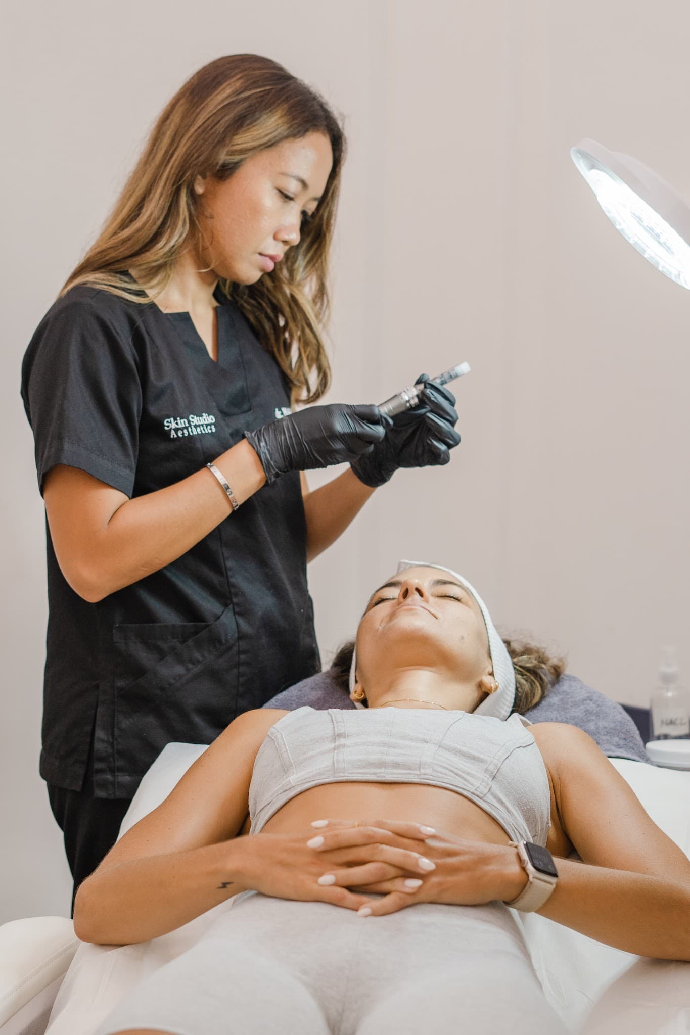 Aesthetician in black branded scrubs preparing a microneedling device while client lies relaxed on treatment bed under a ring light. Both subjects clearly visible, professional setting, warm tones, and excellent composition showcasing the service experience.