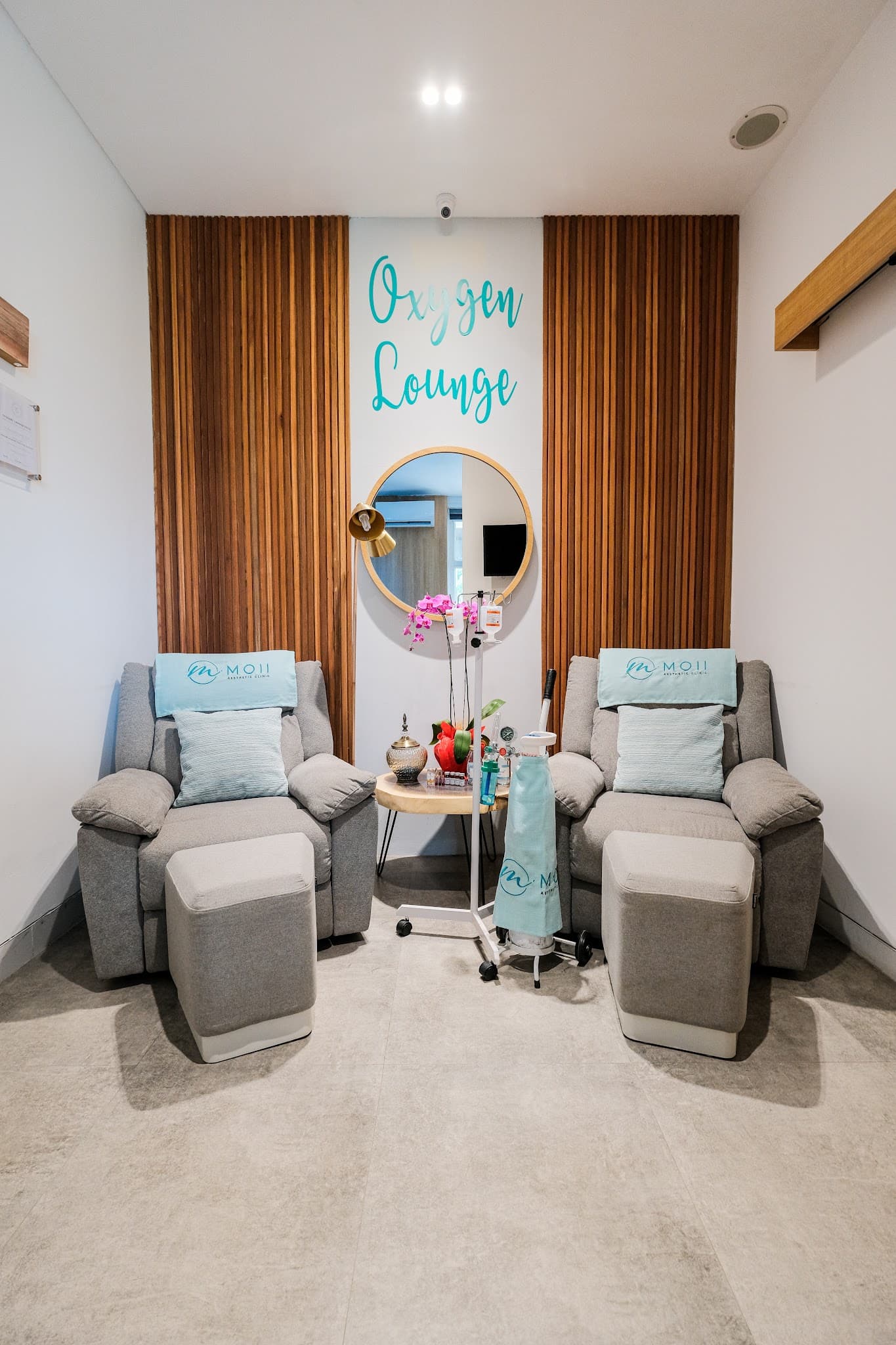 Well-composed, professionally photographed oxygen lounge featuring two plush grey recliners with branded pillows, wood slat paneling, a round mirror, and clean symmetrical framing.