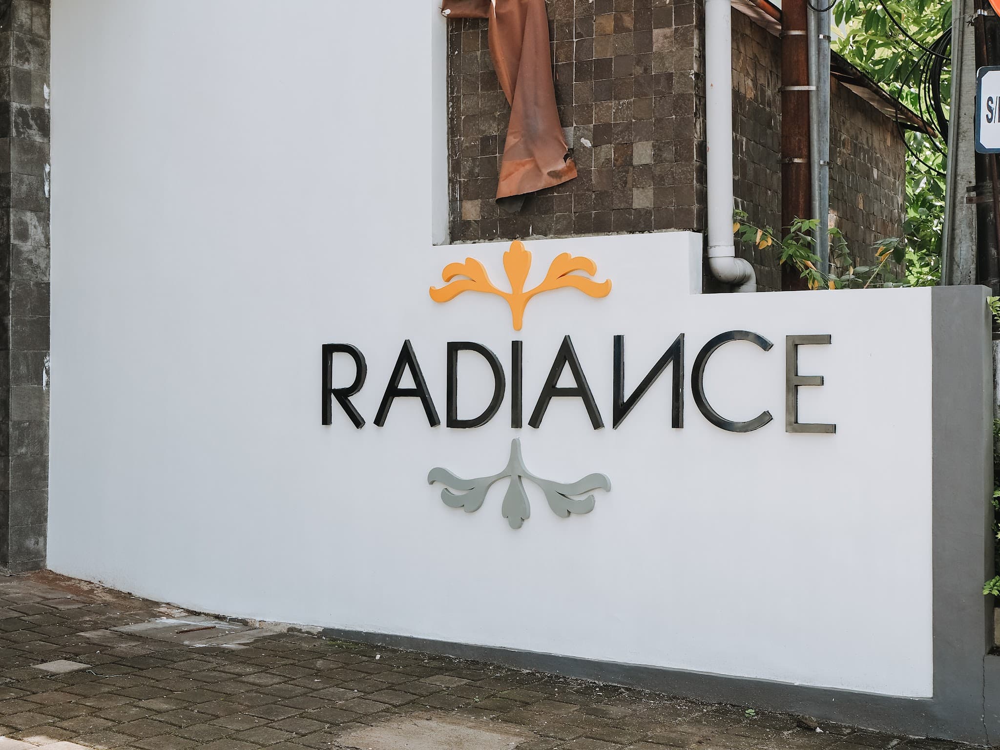 Exterior wall signage of a clinic named 'Radiance' with decorative floral motifs on a clean white wall, though the composition is tightly cropped and includes distracting elements like a draped cloth and exposed plumbing.