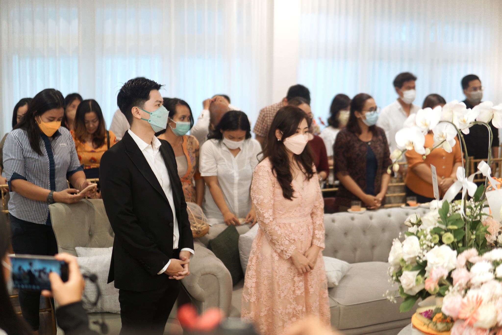 Event or ceremony photo inside the clinic showing a crowd of attendees wearing face masks, with a well-dressed couple in the foreground. Flowers and clinic interior visible in background. Candid moment with human warmth, though masks obscure faces and the image feels more like an event photo than a polished clinic directory image.