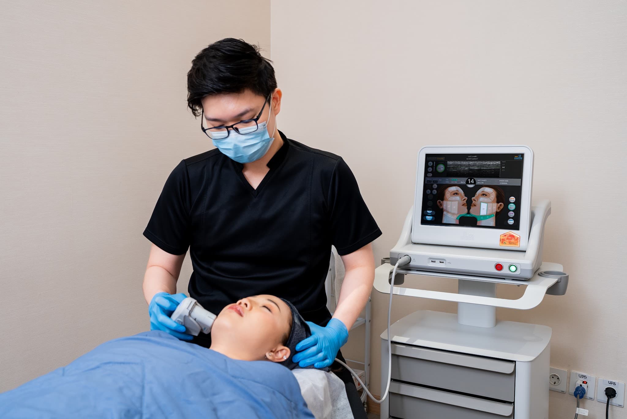 Practitioner in black scrubs performing HIFU facial treatment on relaxed client. Professional equipment visible. Authentic clinical setting with genuine human interaction.