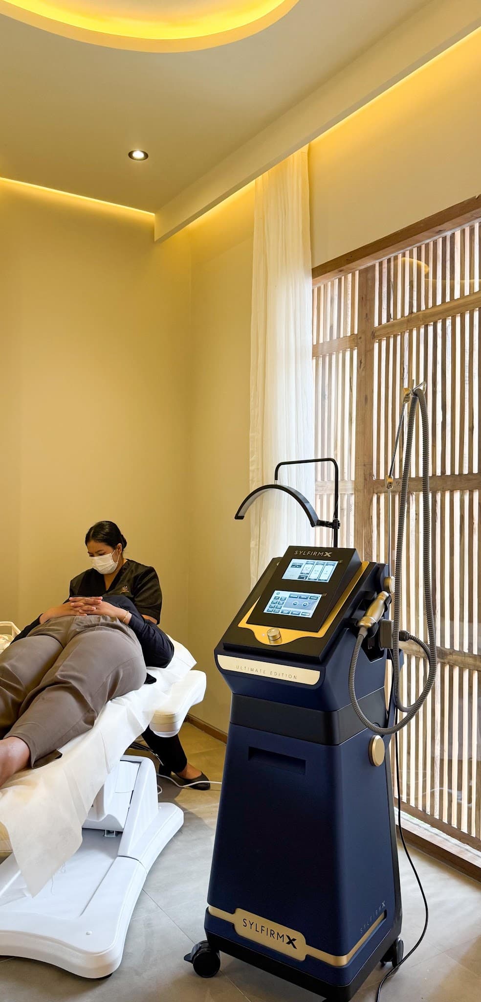 Treatment room showing practitioner performing facial treatment on client with Sylfirm X device. Warm ambient lighting with golden LED ceiling accents and wooden slatted window detail create a premium atmosphere.