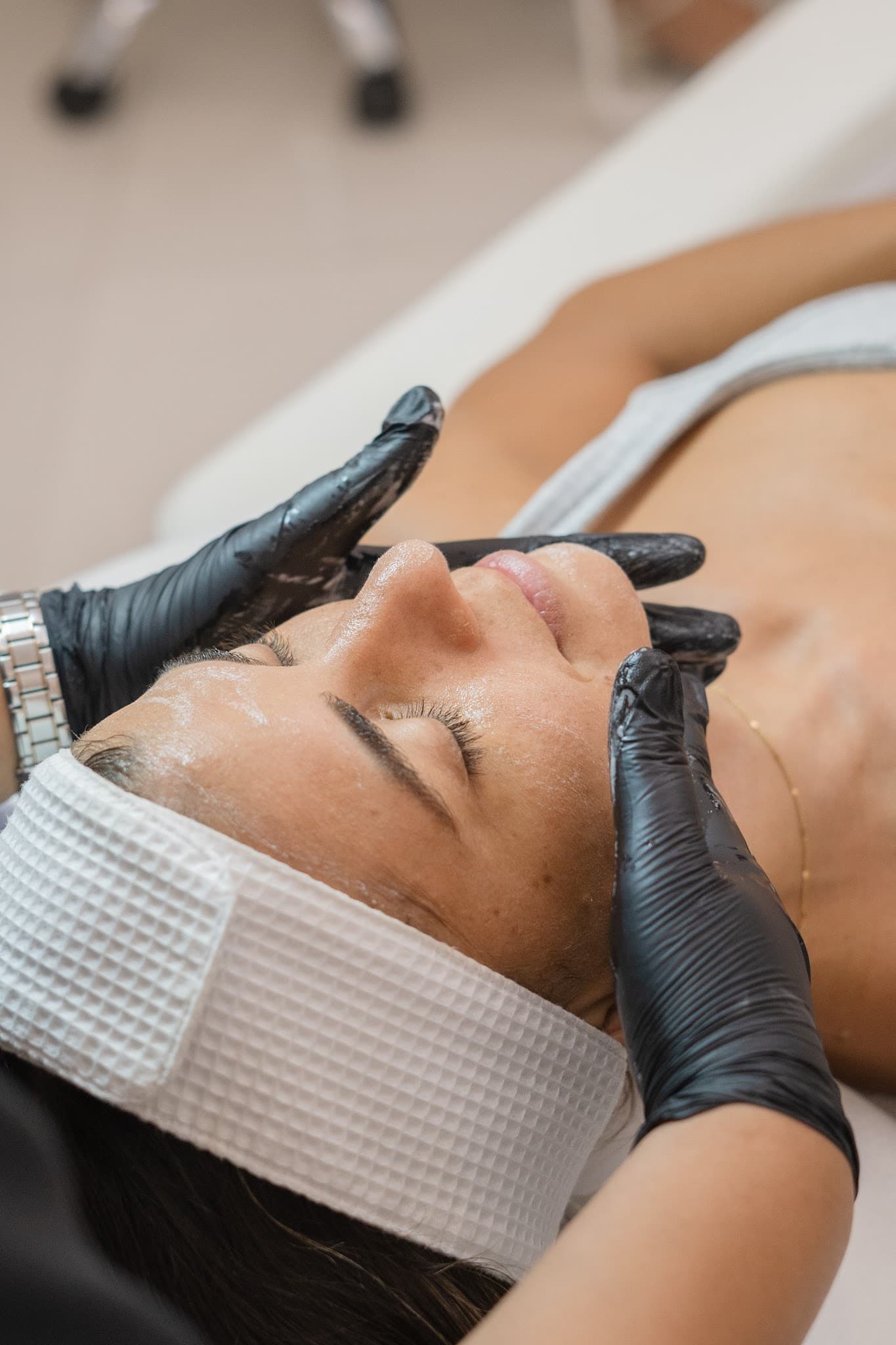Close-up of a facial treatment in progress with practitioner's gloved hands gently working on a client's face. Client is relaxed with eyes closed, wearing a white headband. Professional, warm, and well-lit image showcasing hands-on skincare service.