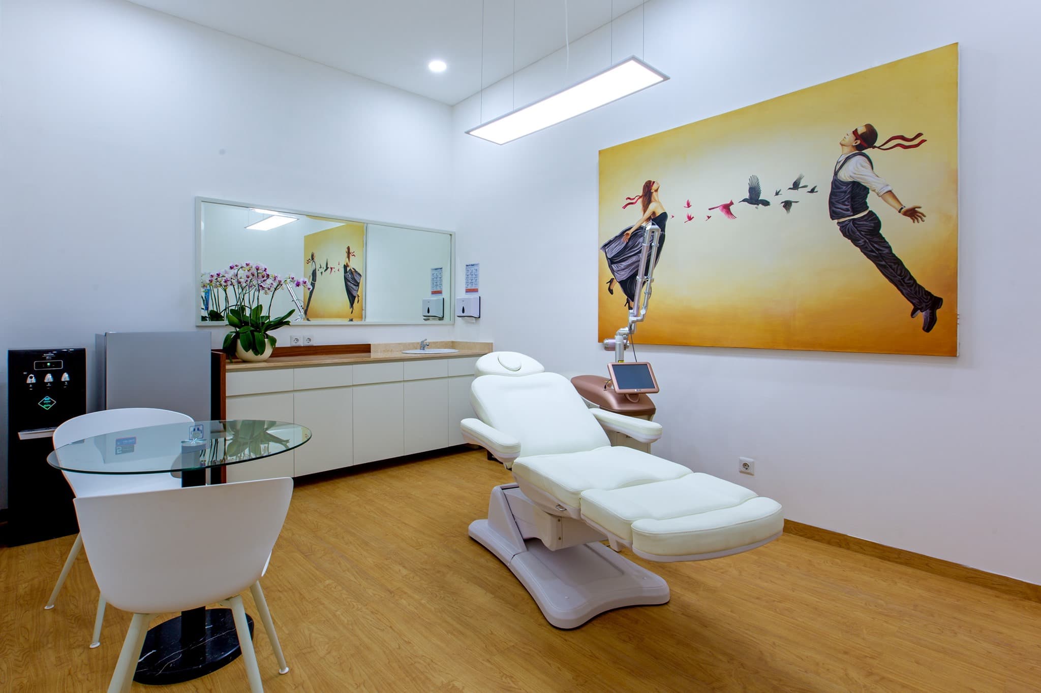 Immaculate treatment room with a white treatment chair, aesthetic equipment, warm wood flooring, elegant consultation area, and a large vibrant art piece creating a luxurious atmosphere.