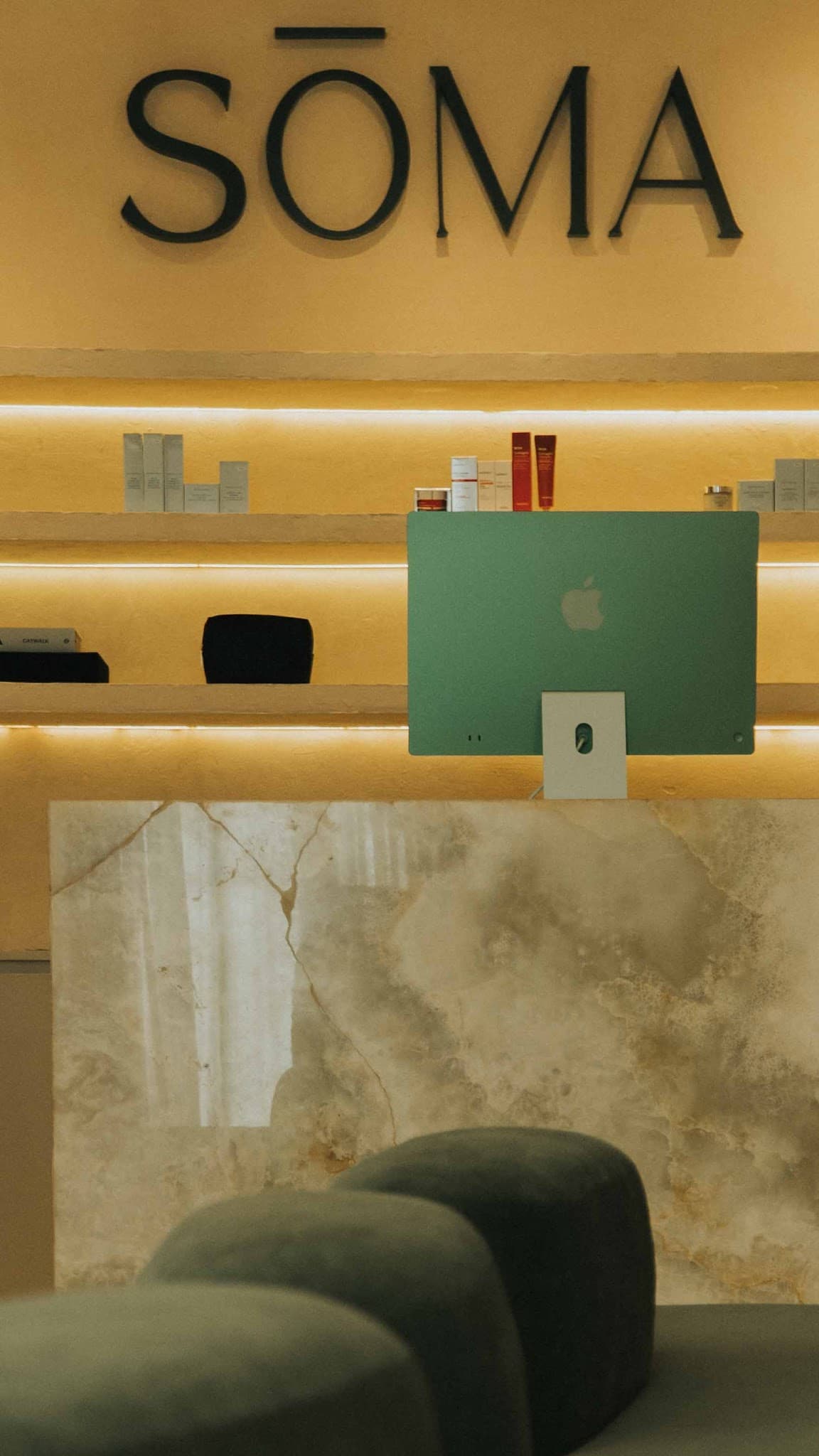 A stylish reception area featuring the SOMA brand signage, warm LED-lit product shelves, a marble counter with a green iMac, creating a modern premium clinic atmosphere.