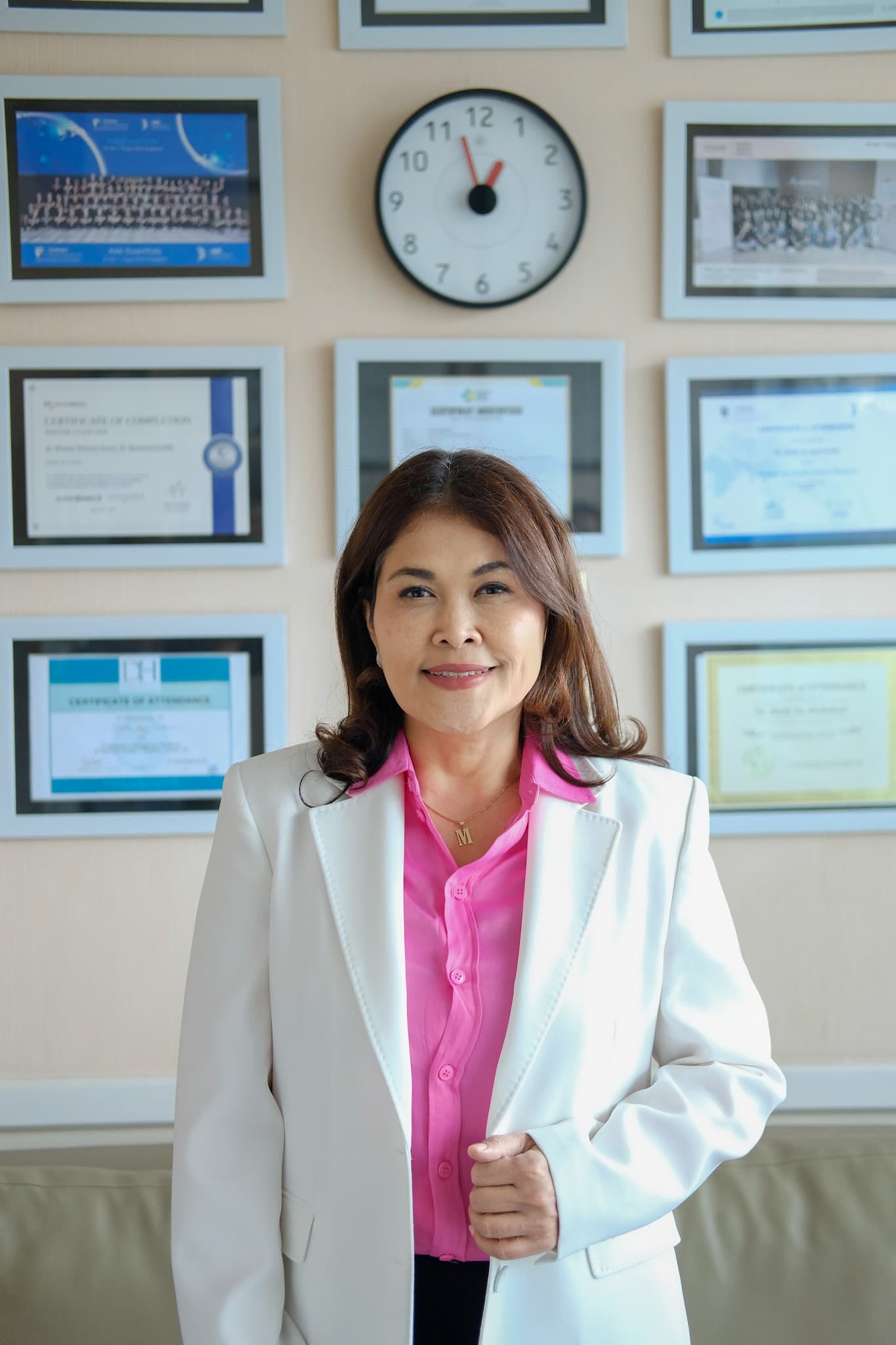 Professional portrait of a female doctor in a pink blouse and white blazer standing before a wall of framed certificates. Gentle smile and professional posture convey trustworthiness.