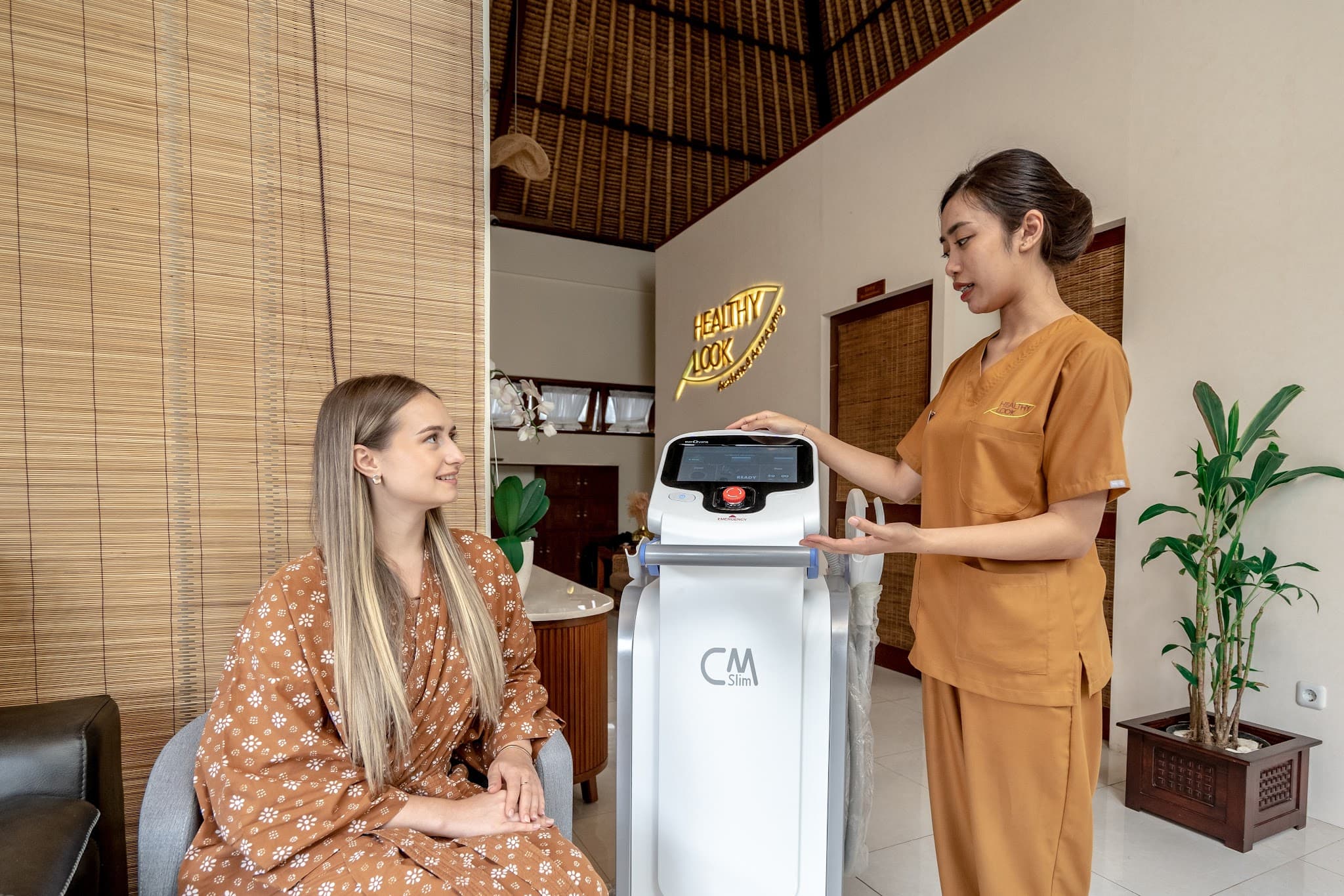 Staff member in mustard uniform presenting a CM Slim body contouring machine to a smiling client in the clinic's Balinese-style reception area with thatched ceiling, bamboo blinds, and the Healthy Look illuminated sign. Warm, inviting atmosphere with excellent professional photography.