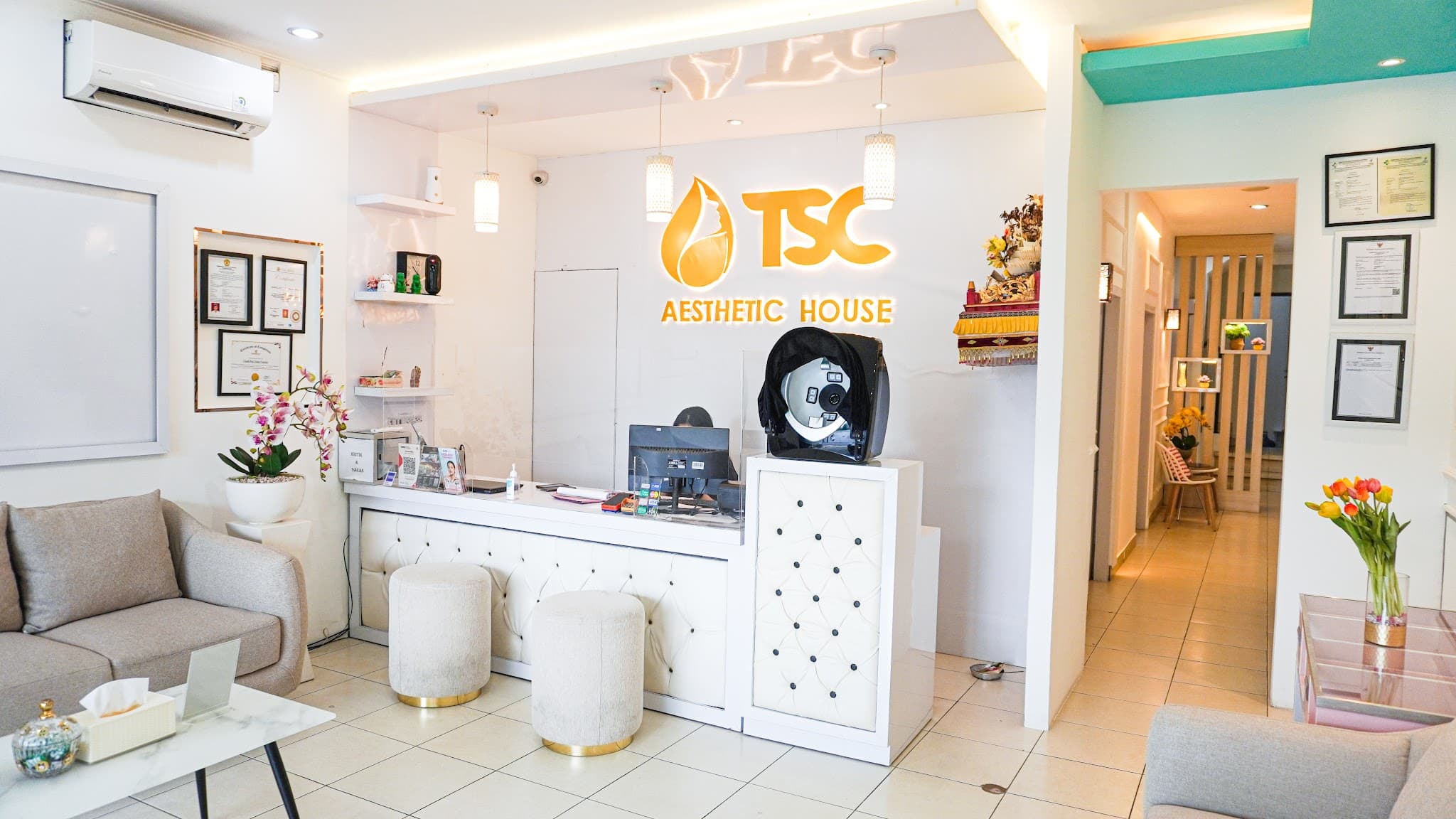 Reception area with prominent TSC Aesthetic House branding, white tufted front desk, and comfortable seating, though the space feels somewhat busy with visible certificates, equipment, and a shrine.