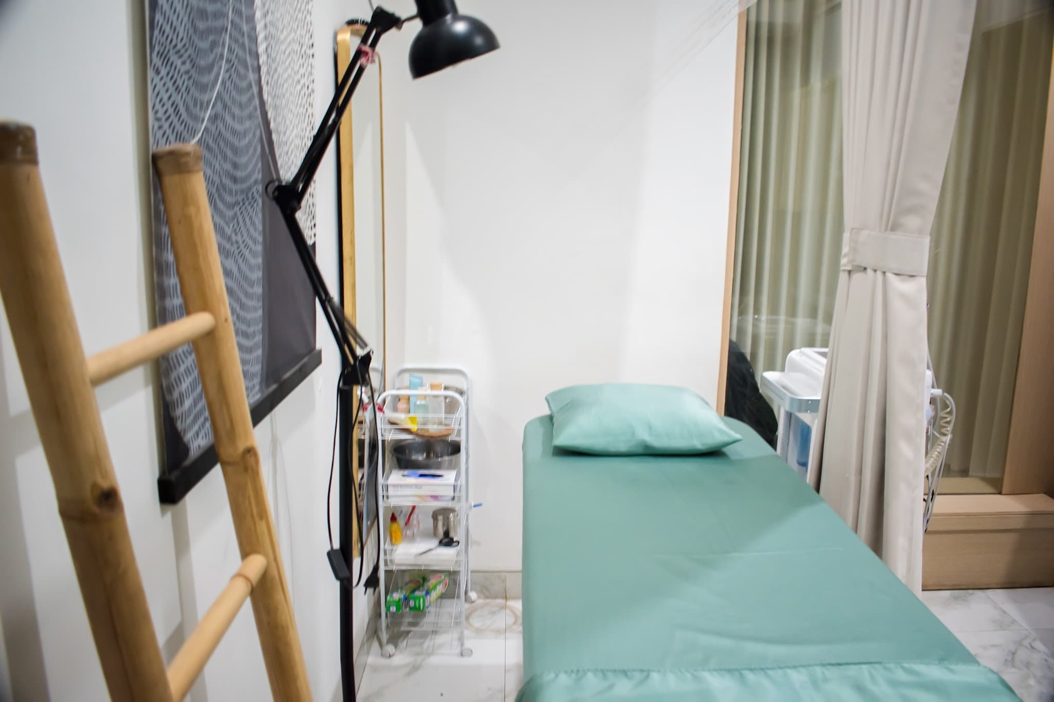 Treatment room with a teal-sheeted bed, magnifying lamp, small supply cart with visible clutter, curtain divider, and bamboo decorative ladder.