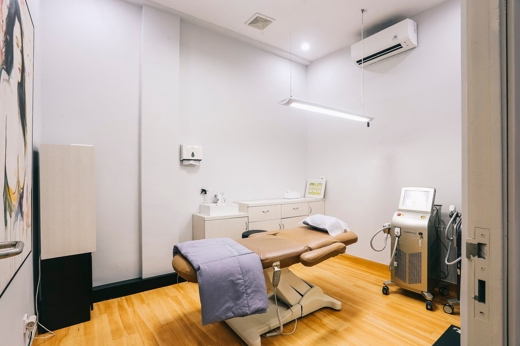 Clean treatment room with tan leather bed, laser equipment, wooden flooring, wall-mounted AC and cabinetry. Well-lit professional space.