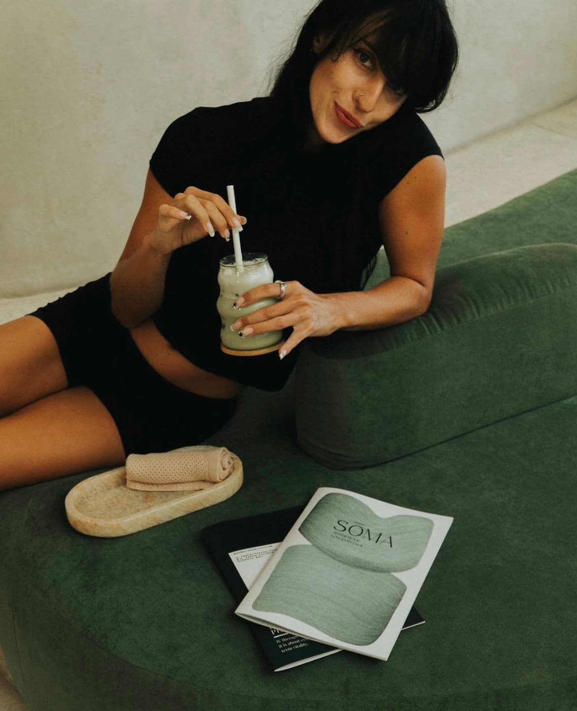 A client relaxes on a plush green velvet sofa with a drink and a SOMA clinic brochure, conveying a premium lounge atmosphere with human warmth.
