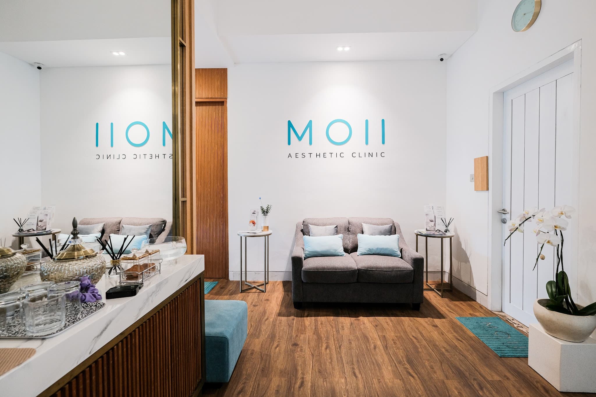 Modern and inviting clinic reception area with clean white walls, branded signage, comfortable grey sofa, warm wood flooring, and elegant decor including orchids and a mirrored accent wall.