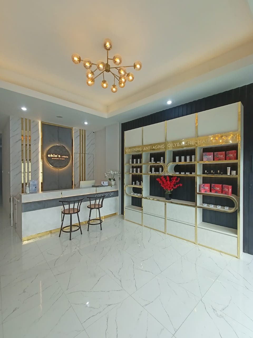 Elegant full view of clinic reception with branded desk, gold-accented product display wall, sputnik chandelier, marble flooring.