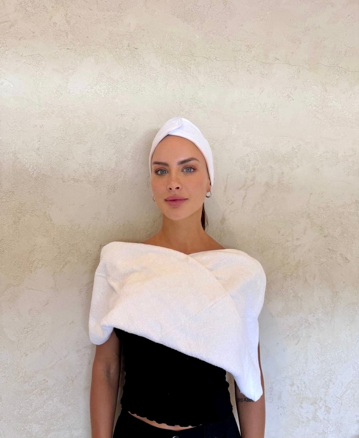 A client poses against a textured beige wall wearing a white towel wrap and headband, projecting a fresh post-treatment glow with a clean aesthetic.