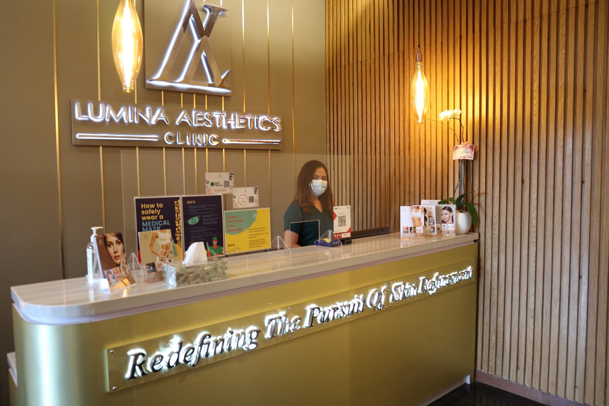 Reception desk of Lumina Aesthetics Clinic with branded signage, warm pendant lighting, wooden slat accent wall, and a receptionist behind a plexiglass barrier with the clinic tagline illuminated on the front desk