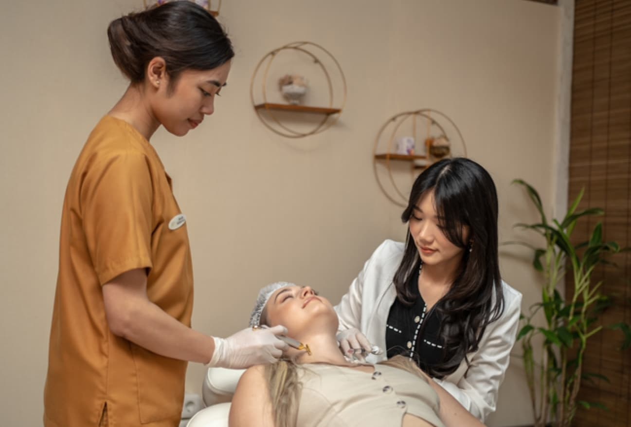 Wider angle of a facial treatment in progress with a doctor and assistant attending to a client reclined on a treatment chair. Warm, professional atmosphere with decorative circular wall shelves and greenery in the background.