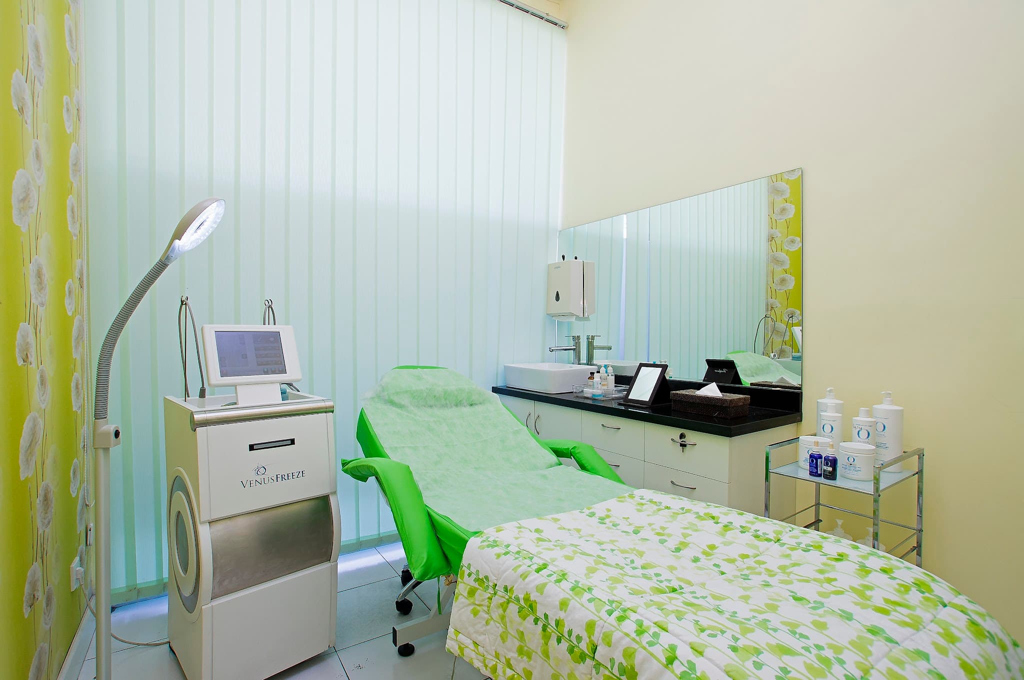 Clean treatment room with green color scheme featuring a Venus Freeze device, treatment chair, vanity area with mirror, and neatly arranged skincare products. Well-lit and organized clinical space.