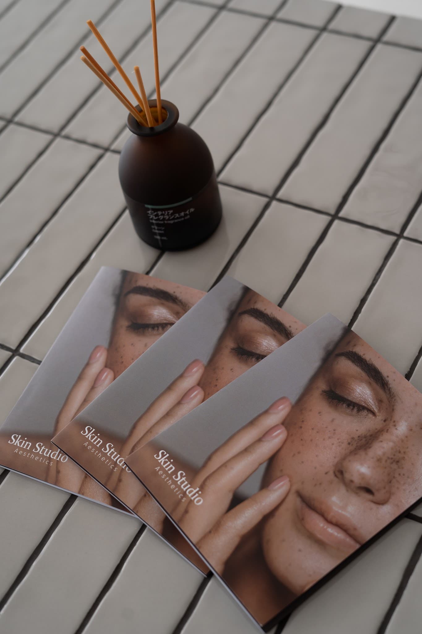 Styled flat-lay of Skin Studio Aesthetics branded brochures fanned out on a tiled surface alongside a reed diffuser. Well-composed detail shot with nice depth of field, though it functions more as branded marketing material than a true interior or service image.