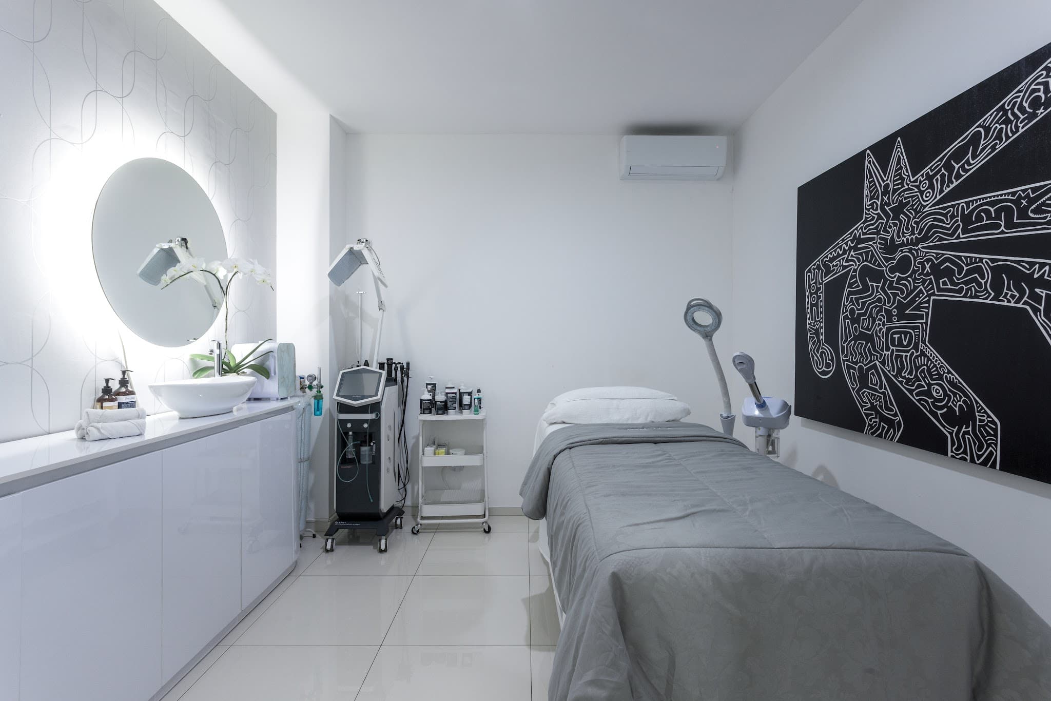 Well-composed treatment room with clean white and grey aesthetic, bold black-and-white mural art, backlit oval mirror, treatment bed with grey linens, professional equipment, magnifying lamp, and orchid accents.