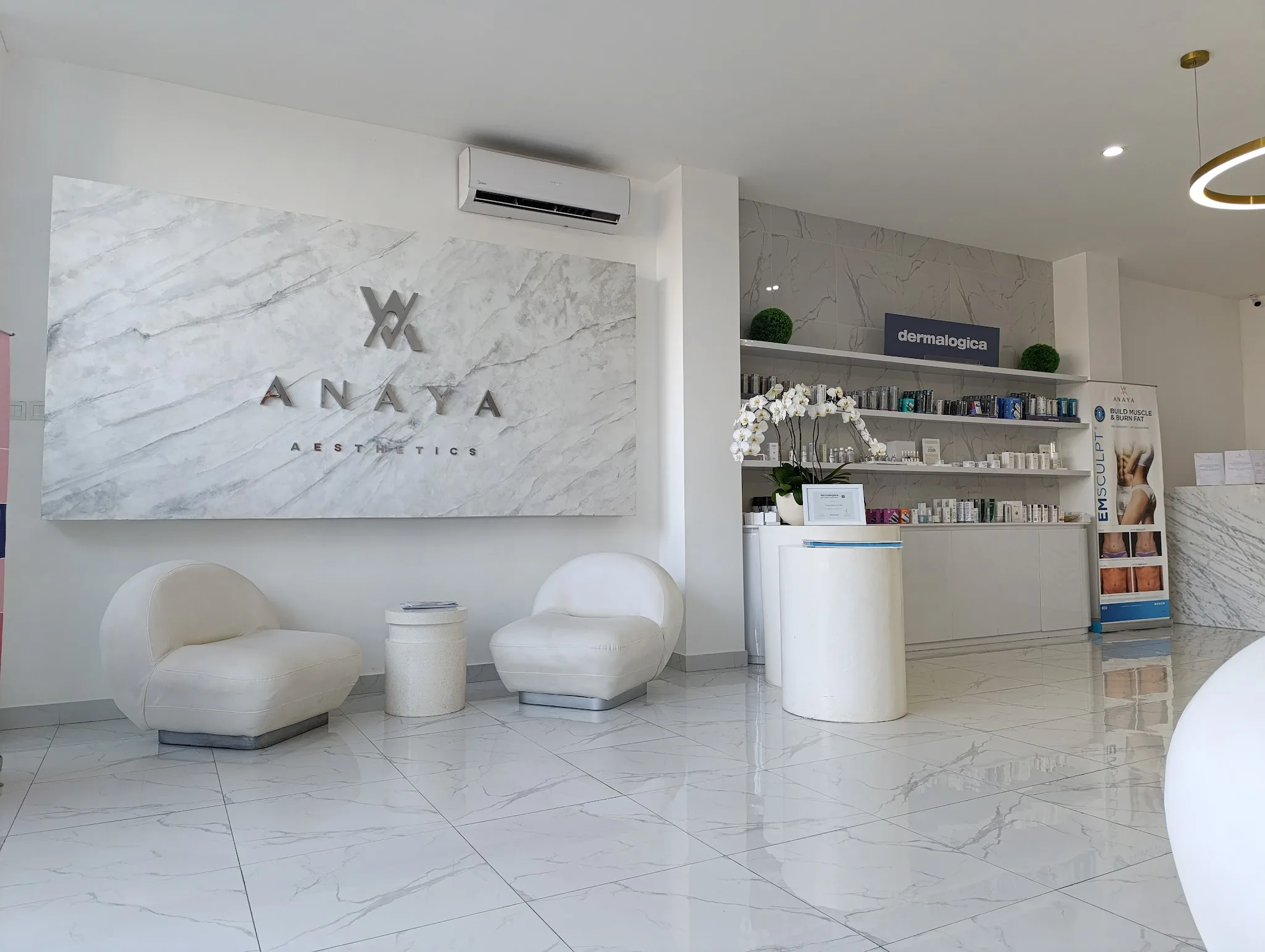 Three smiling staff members posing in front of a marble-clad clinic wall with logo, product shelves visible on the right. Warm and welcoming team photo with good human presence and branded uniforms.