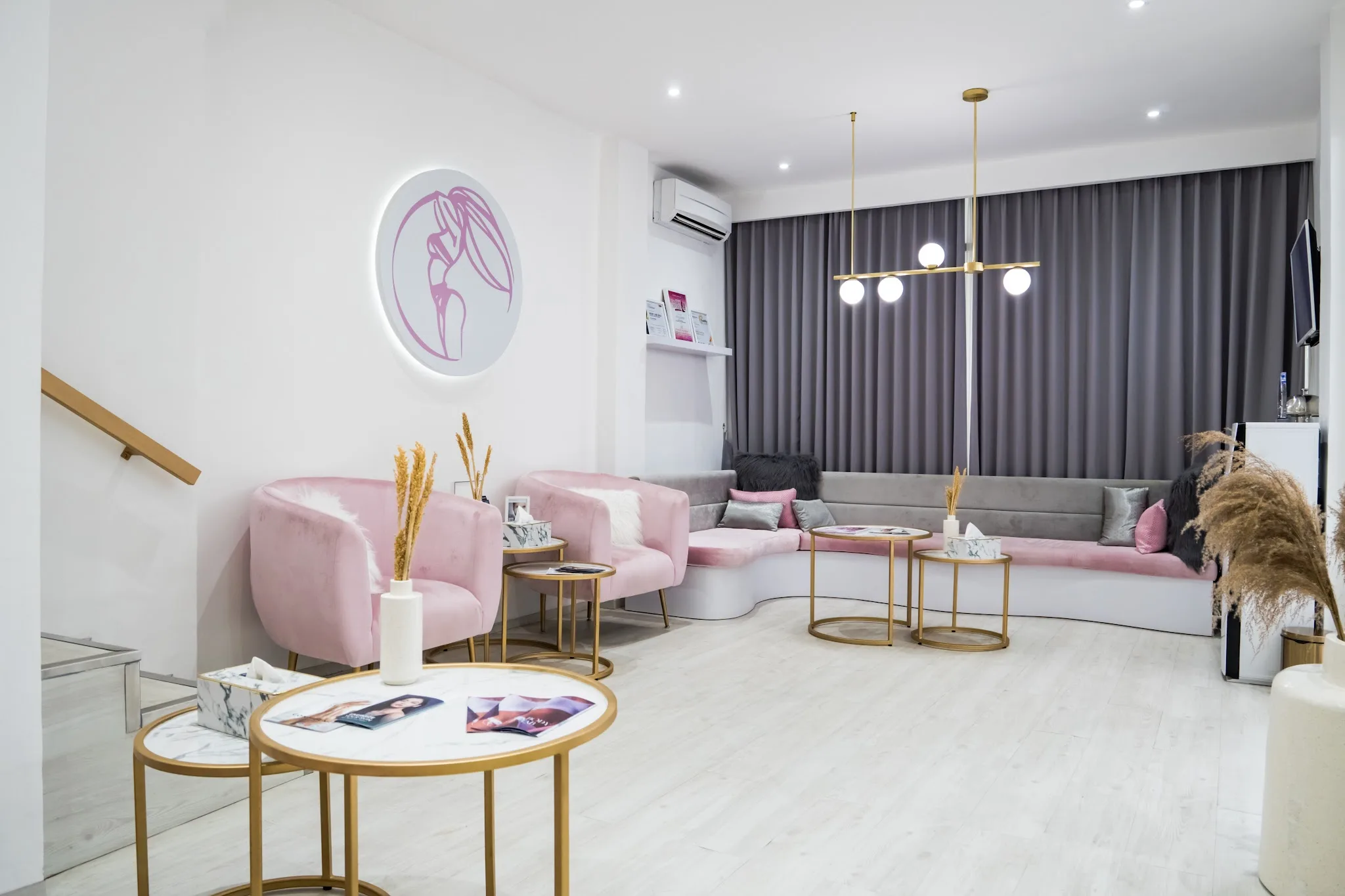 Clinic waiting area with pink velvet armchairs, gold accent tables, decorative staircase with wooden slat divider, and backlit logo sign on wall. Seasonal Christmas decorations and promotional materials are visible.