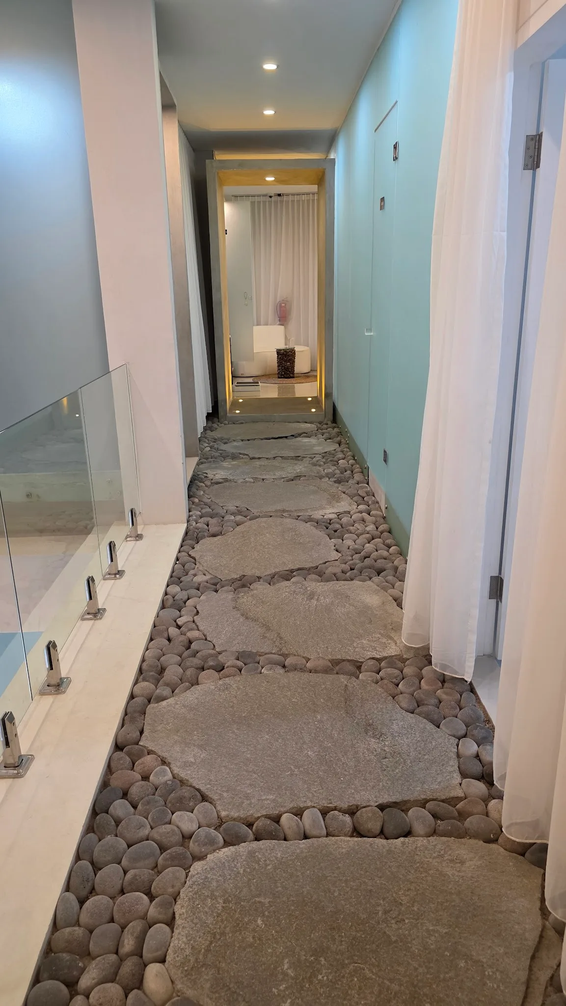 Spa-like hallway corridor with natural stone stepping path over river pebbles, flowing white sheer curtains, concrete wall elements with warm downlighting.