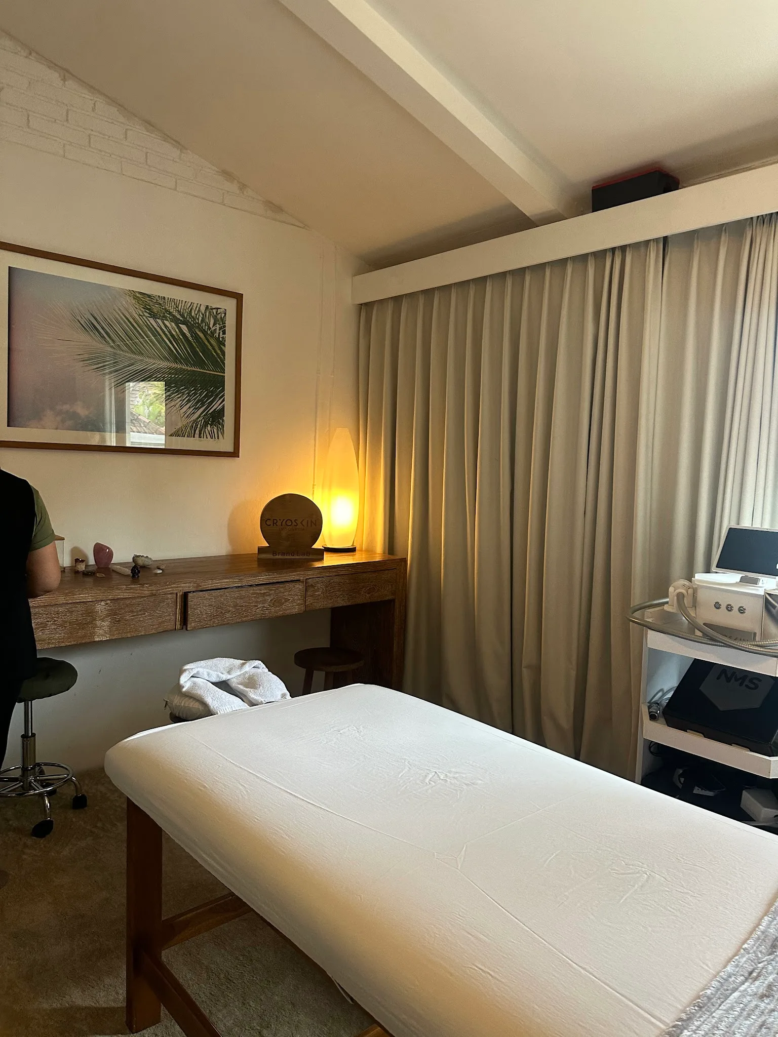 Treatment room with Cryoskin machine, wooden console table, tropical framed artwork, warm ambient lamp, and treatment bed with white linens in a cozy spa-like setting