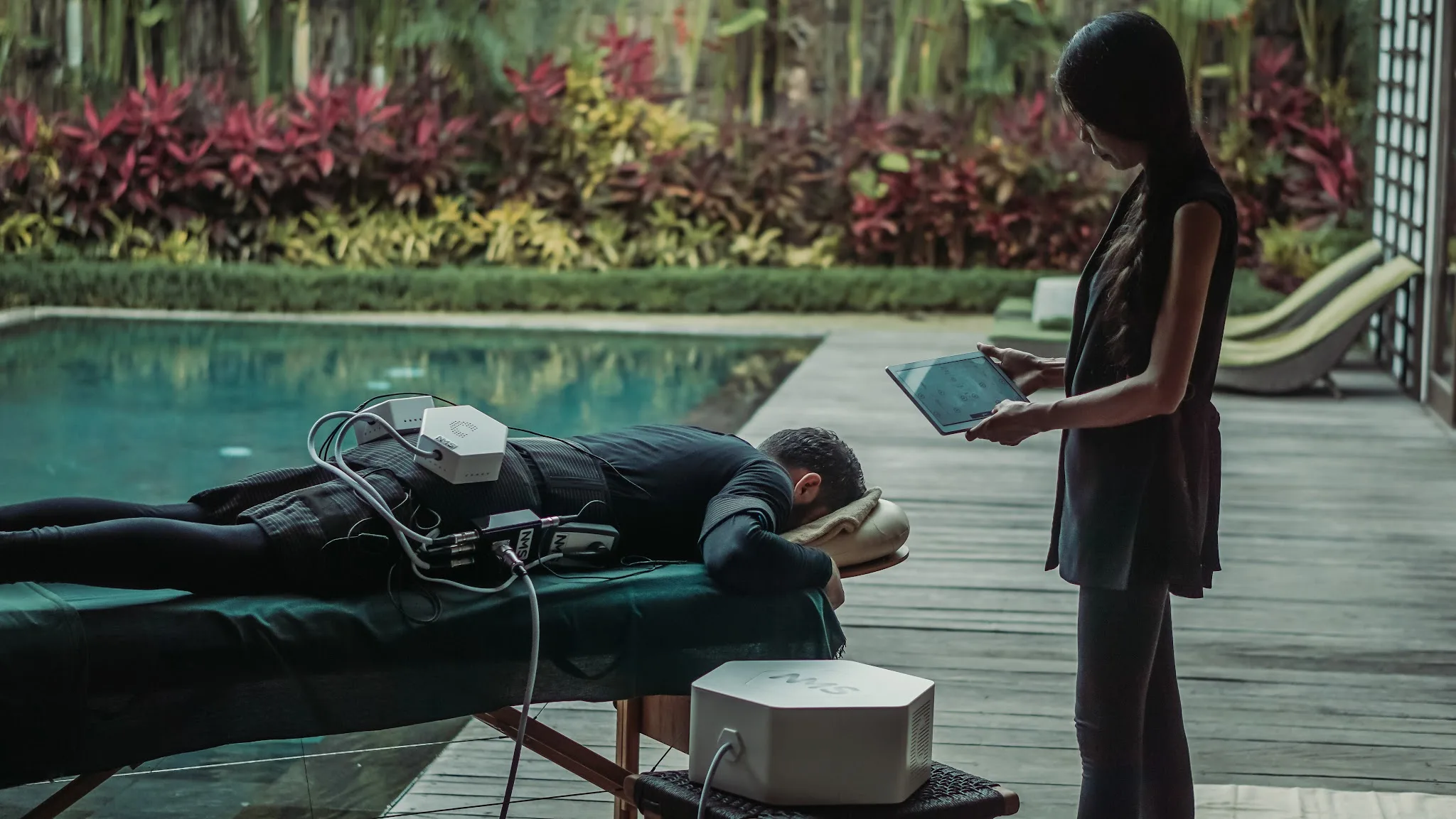 Treatment room showing full massage bed with white sheet, wooden console with Cryoskin signage, warm lamp, tropical artwork, and NMS device cart