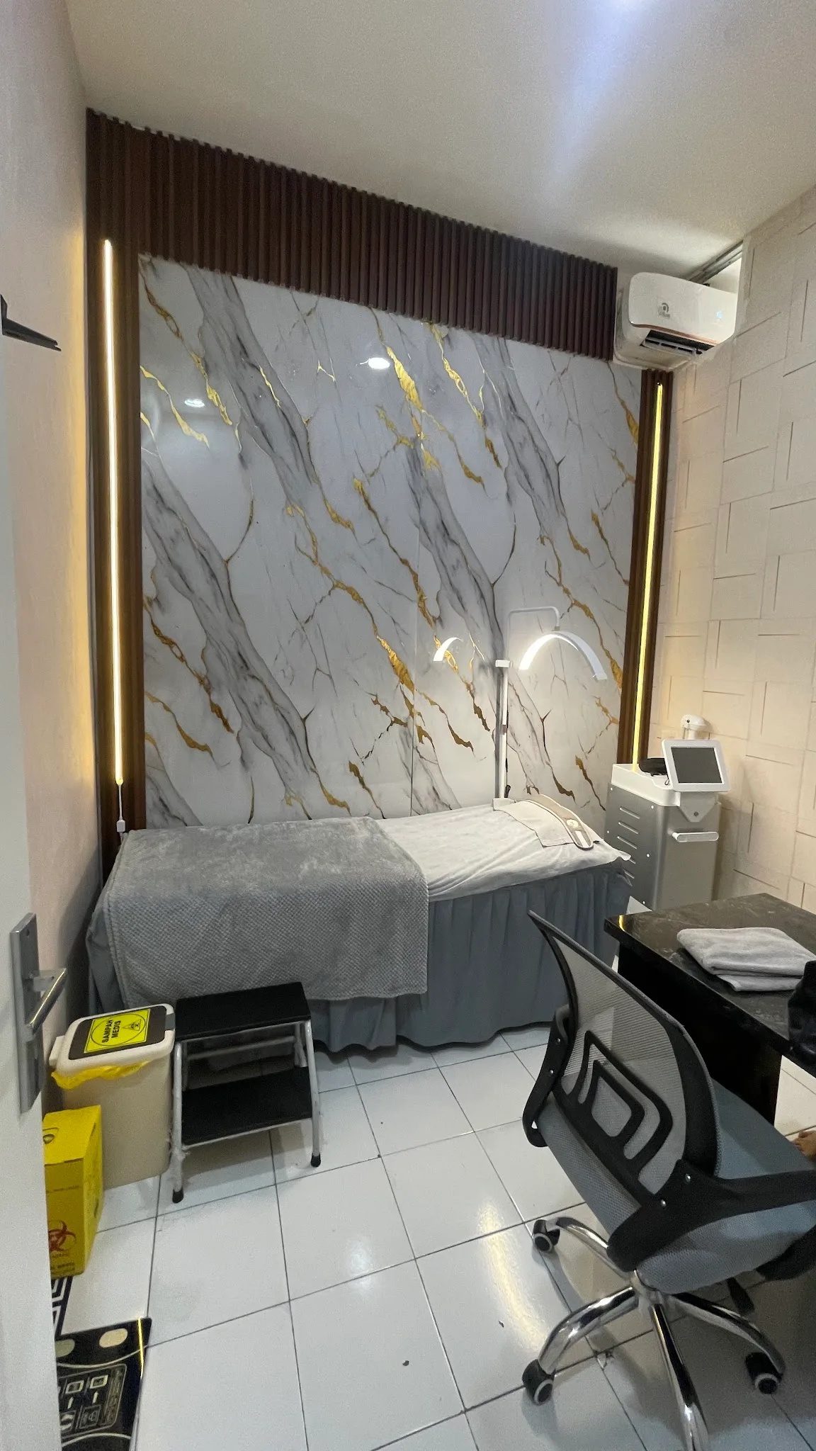 Close-up of single treatment station with VORTA hydrafacial machine, LED face mask, treatment bed, arc lamp against marble walls. Cluttered with visible cables and tissue box.