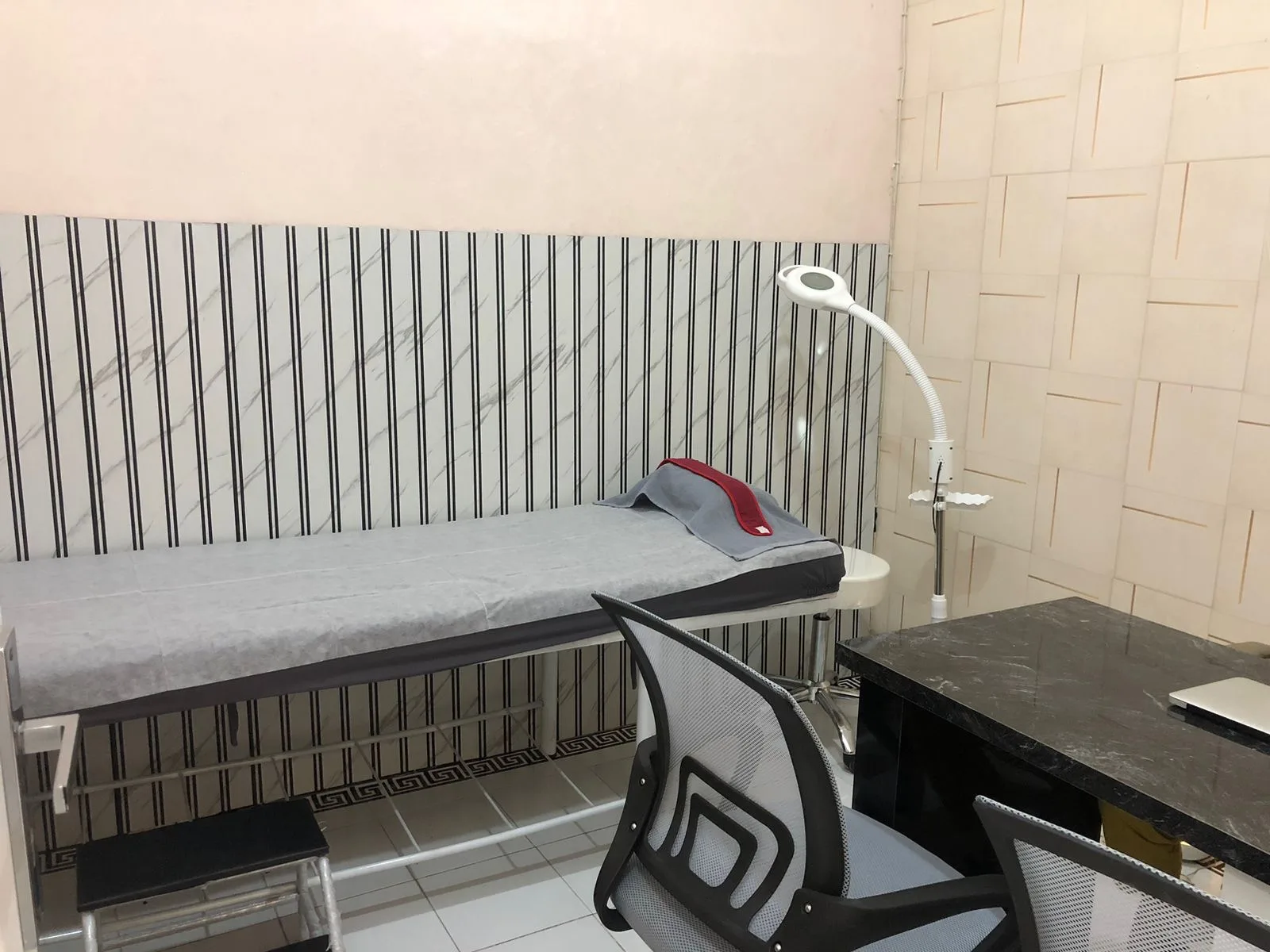 Basic treatment room with striped wallpaper backdrop, examination bed with disposable sheet, magnifying lamp, office chair, and dark marble desk. Modest with harsh lighting, lacking premium aesthetics.