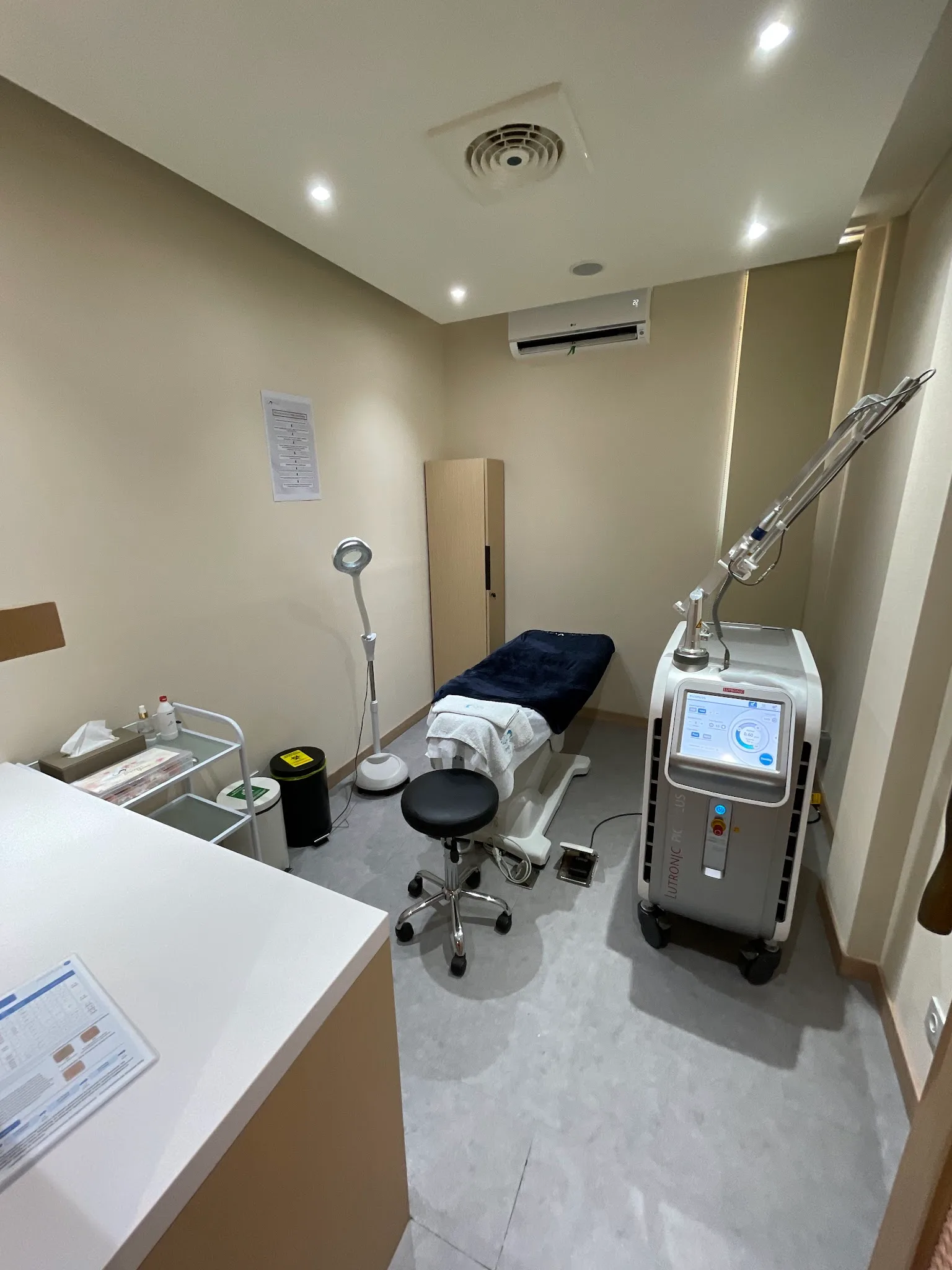 Modern clinic reception area with dark reception desk, staff members, product display shelving with warm perforated wood paneling, monstera plant, comfortable seating. Contemporary premium aesthetic.