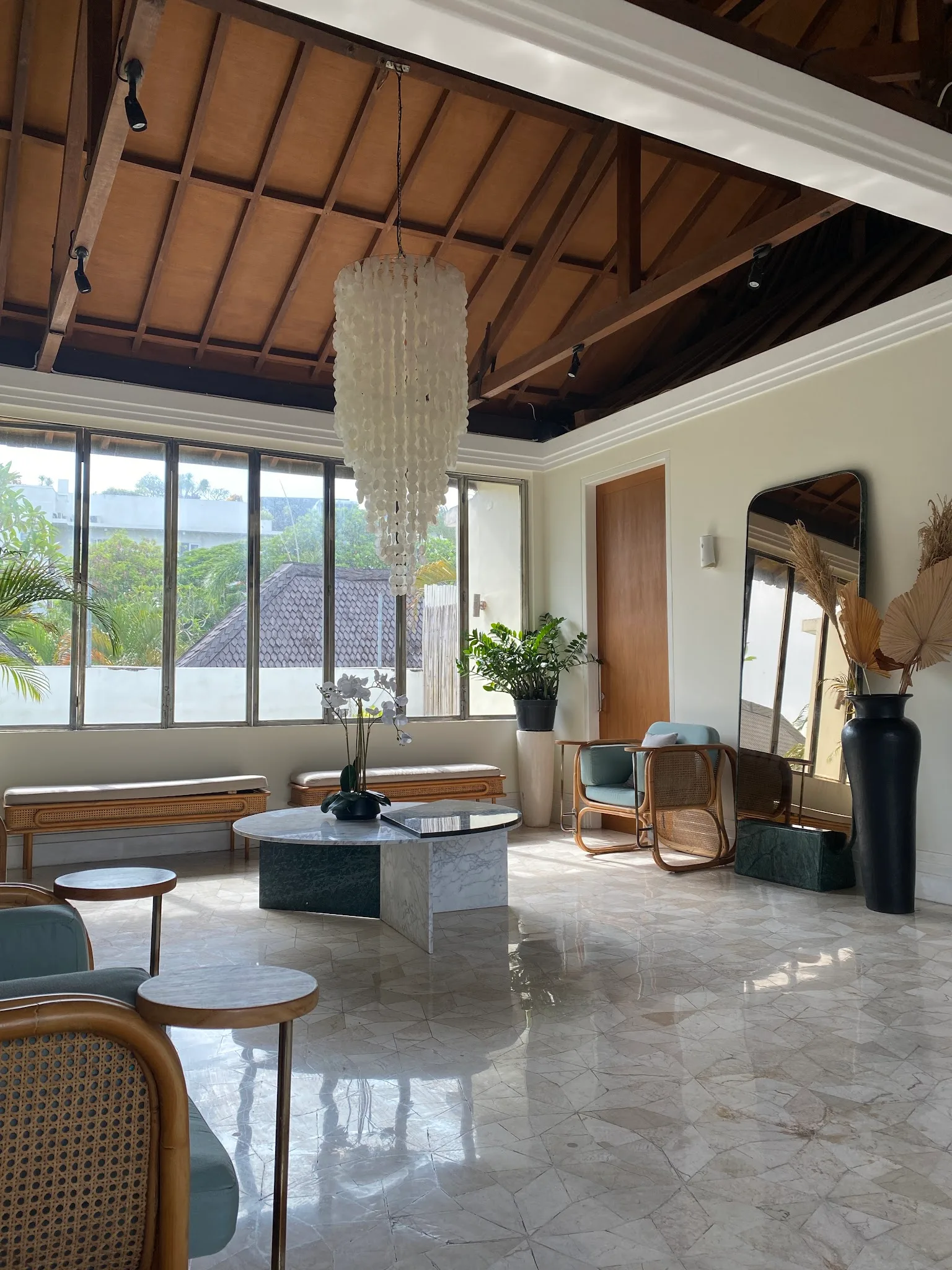 Spacious clinic lobby with marble flooring, rattan-framed chairs with sage upholstery, tropical plants, artwork, and natural light from large windows. A promotional banner on the left detracts from the otherwise elegant space.