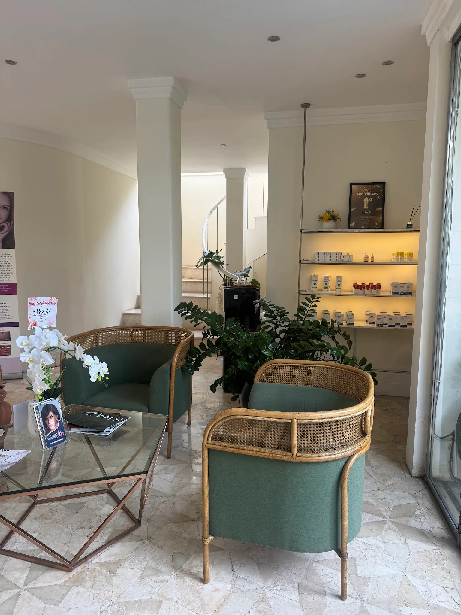 Wide-angle view of clinic interior showing a lounge area with natural light, plants, marble flooring, and seating. A prominent promotional pull-up banner and a QR code price list sign clutter the composition.